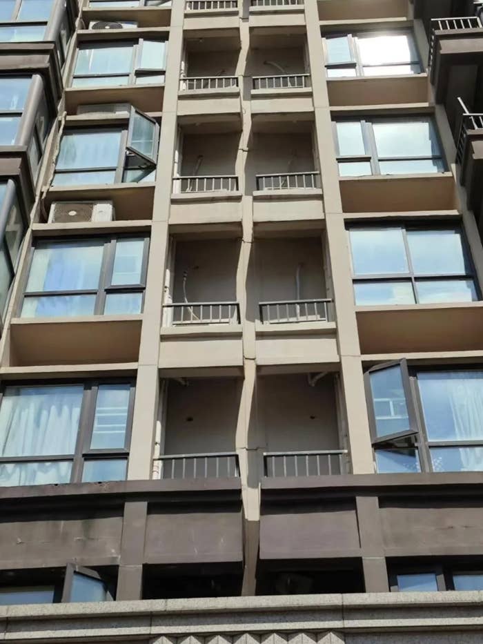 Cracked exterior of a high-rise building, showing structural damage between balconies. Some windows are open
