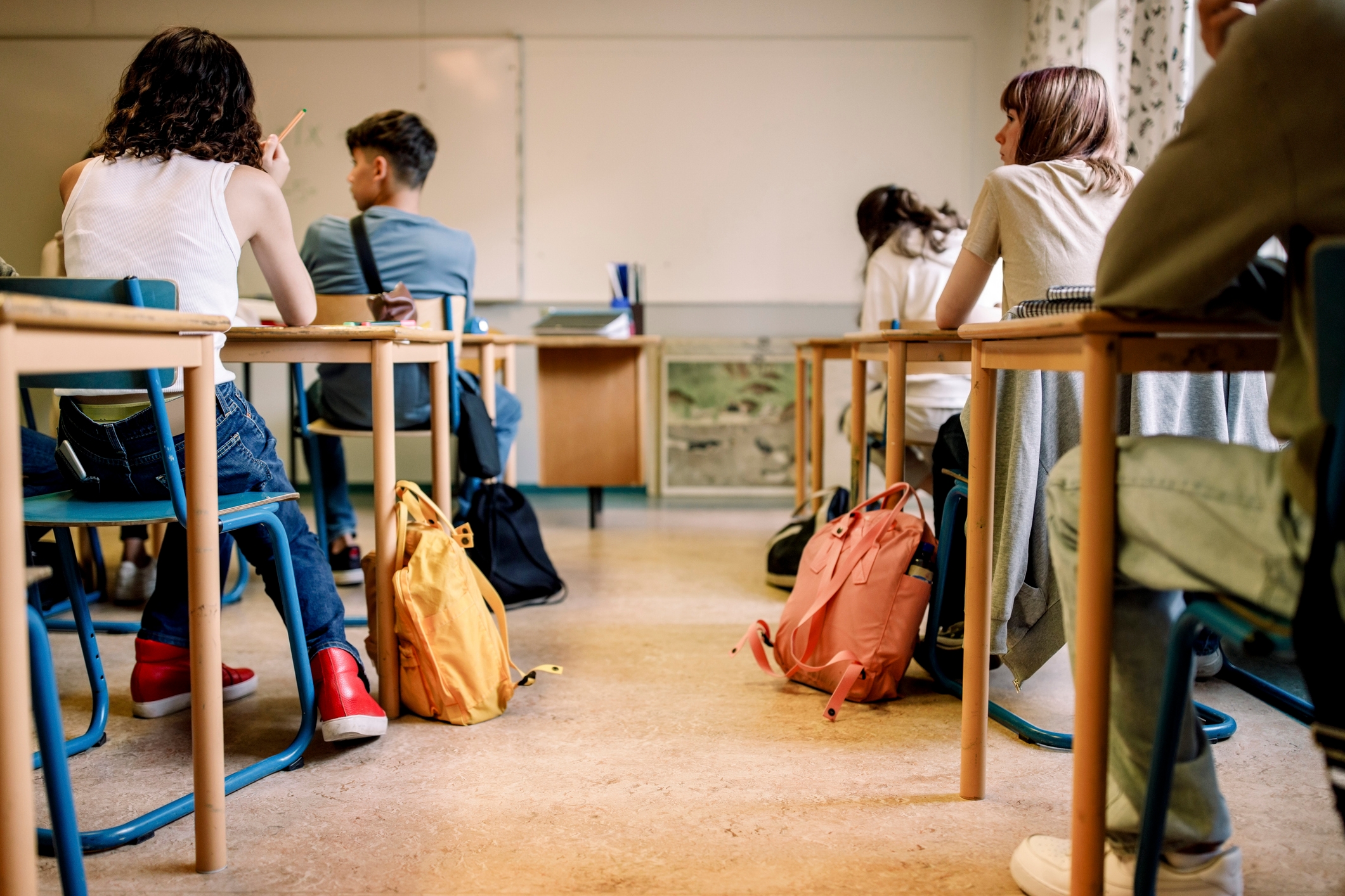 Students sit at desks in a classroom, listening intently to a lesson. Backpacks are on the floor near the desks