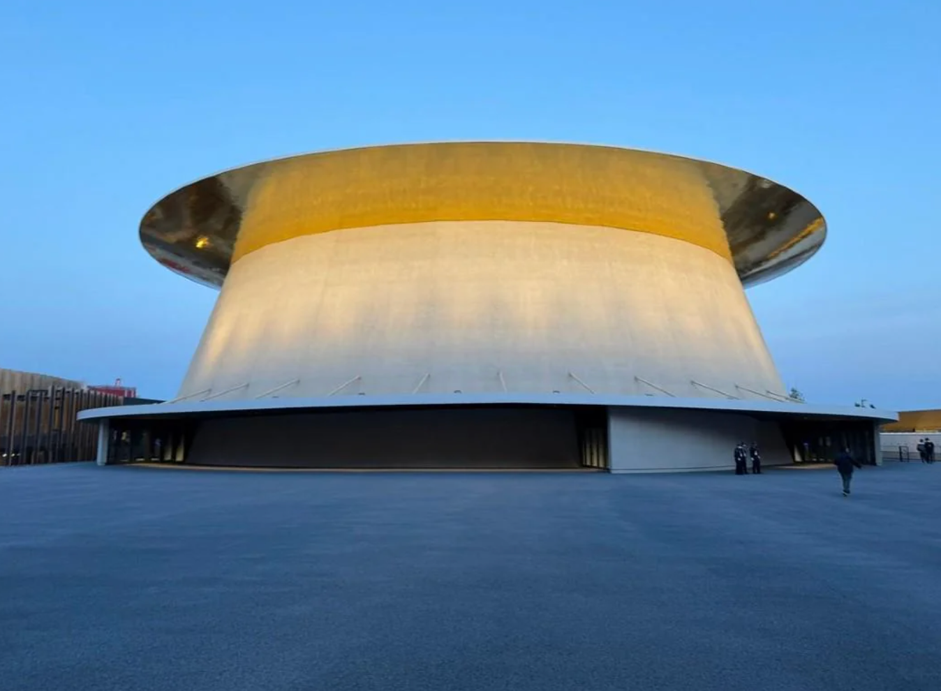 Futuristic, disc-shaped building with reflective surface and wide base, situated in a spacious area under a clear sky