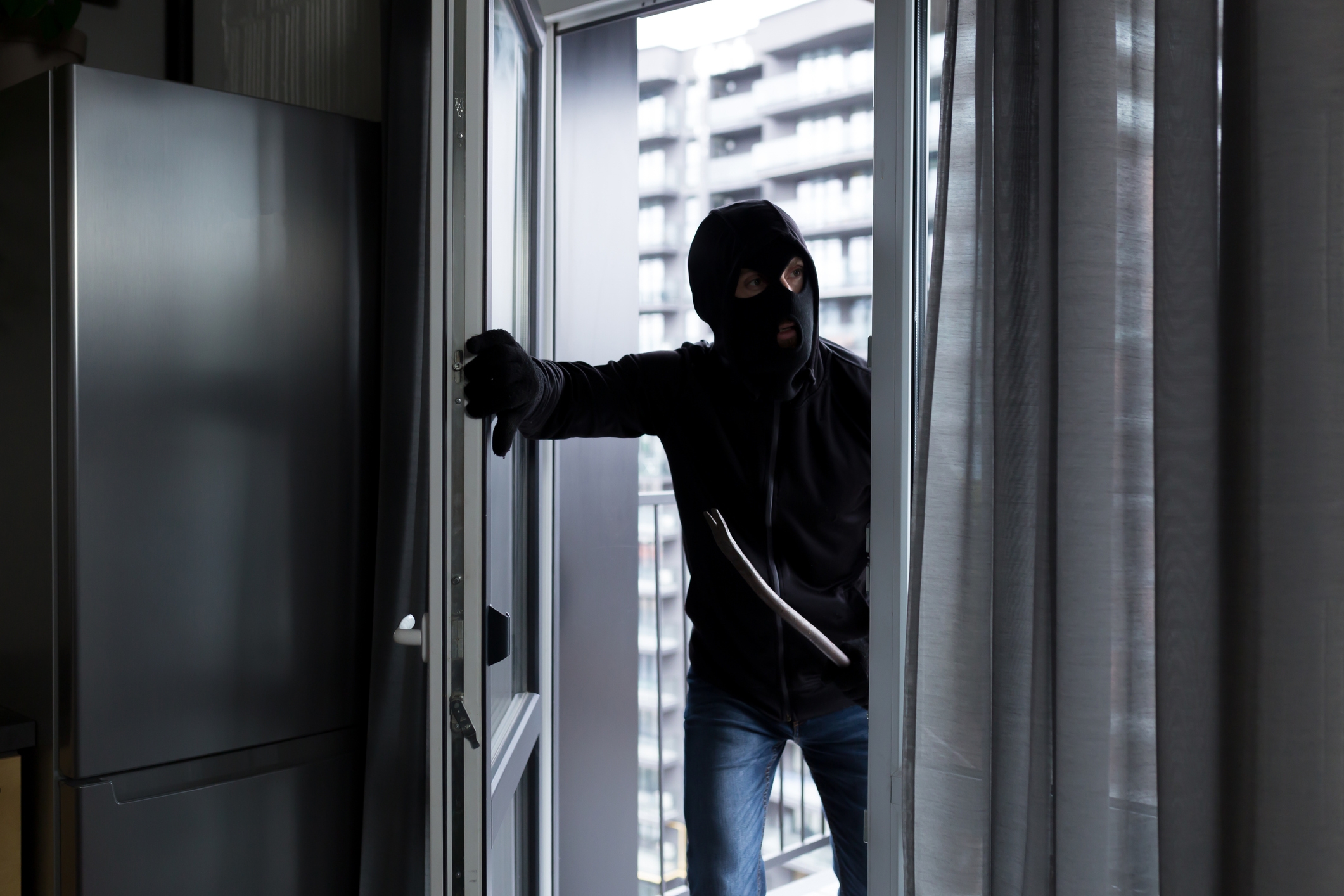 Person in a balaclava and gloves entering through a balcony door, suggesting a burglar or intruder scene