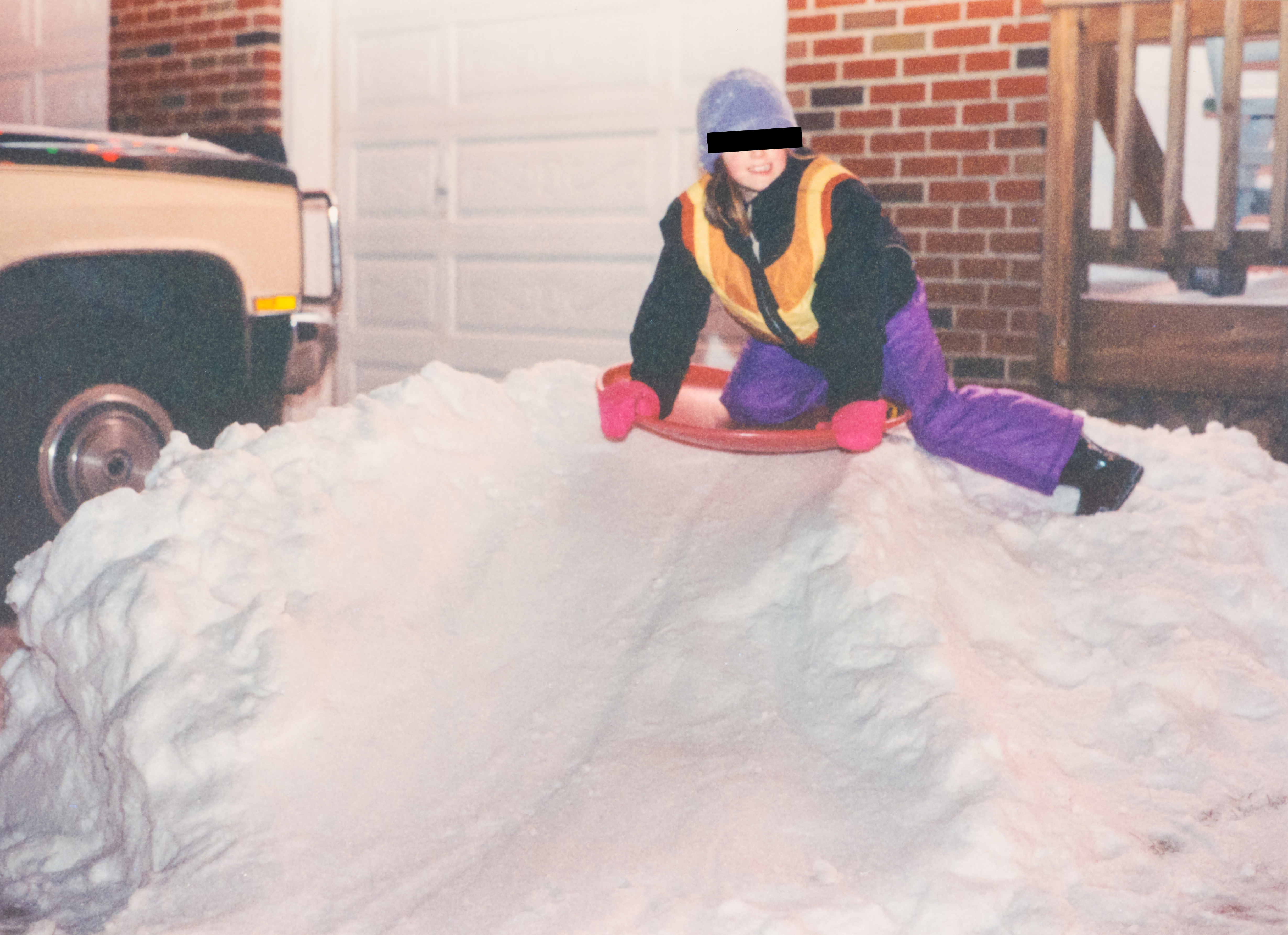 Child in winter gear sits on a sled atop a snow mound, ready to slide down. Brick building and vehicle in the background