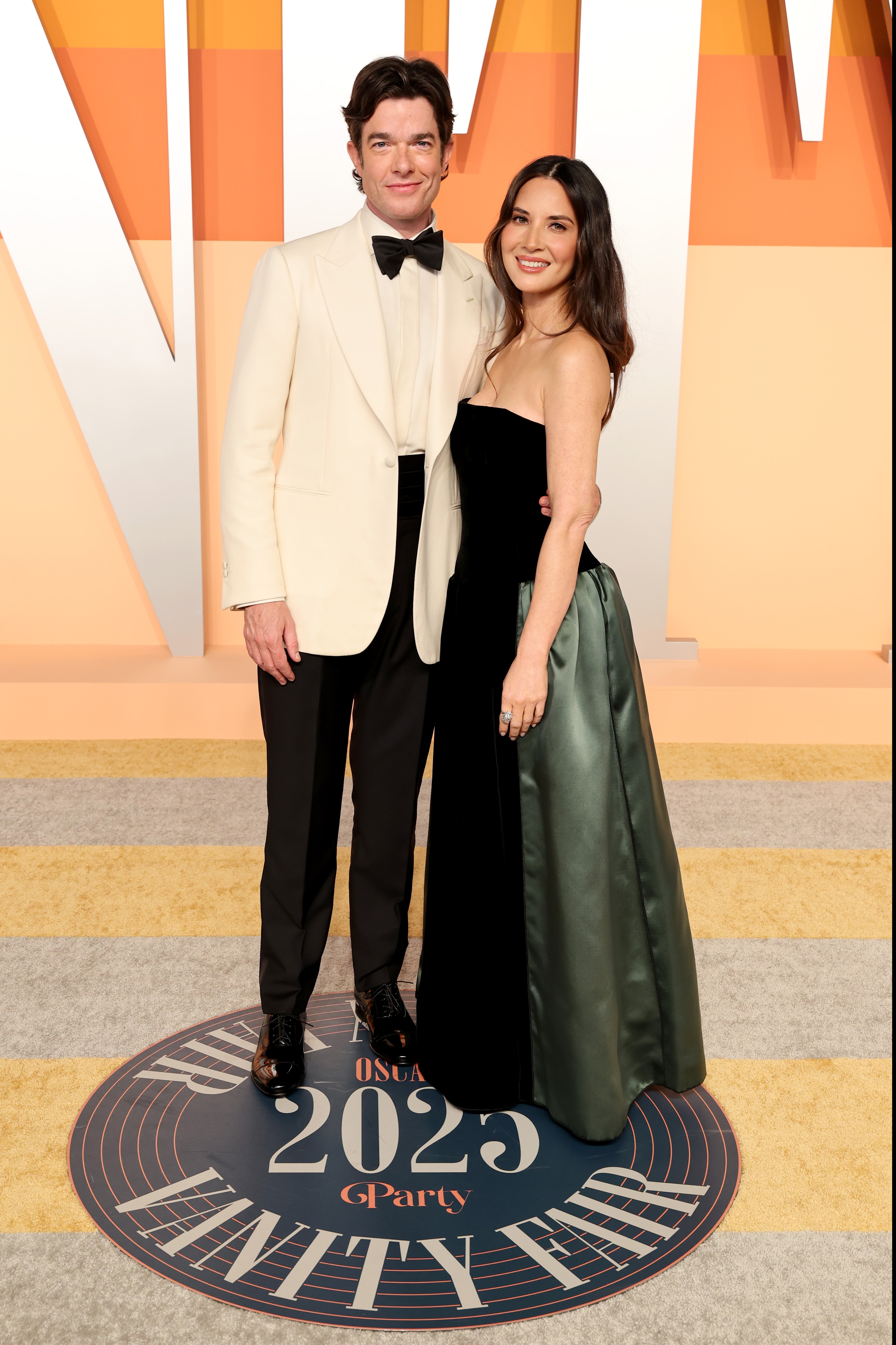 A couple poses on a celebratory event's carpet. The man wears a white tuxedo jacket, and the woman wears a strapless gown