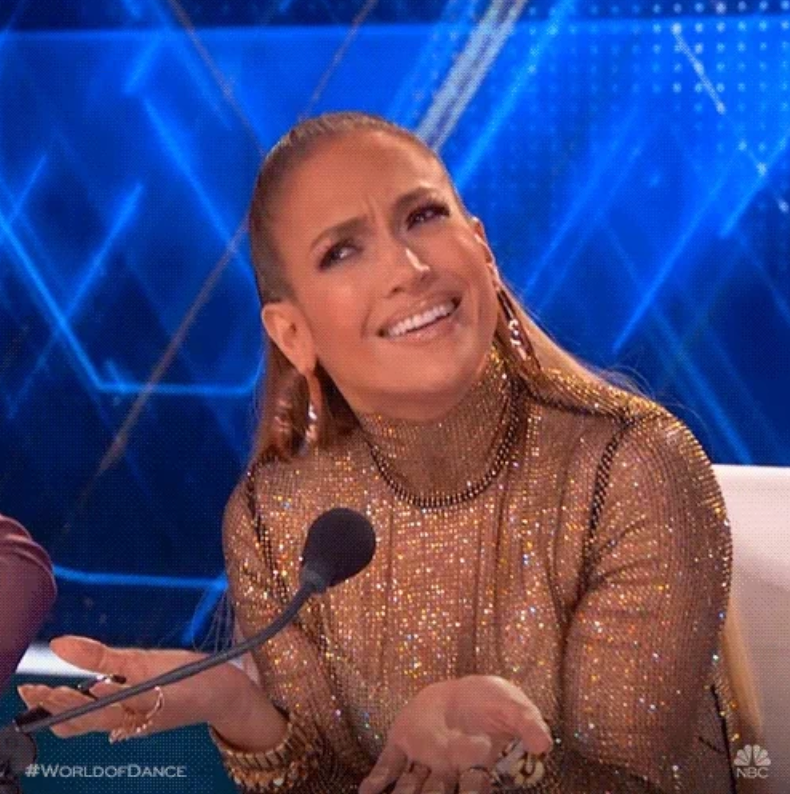 Person gesturing with raised eyebrows and palms up while sitting at a table with a microphone. They're wearing a glittery turtleneck top