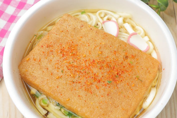 うどんの上に揚げがのった料理。トッピングに七味唐辛子が振られている。背景にピンクのチェック柄の布と葉がある。