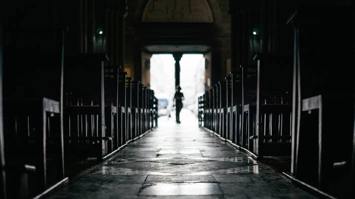 Person walking down a dimly lit church aisle with wooden pews on either side, illuminated by light from an open door in the distance