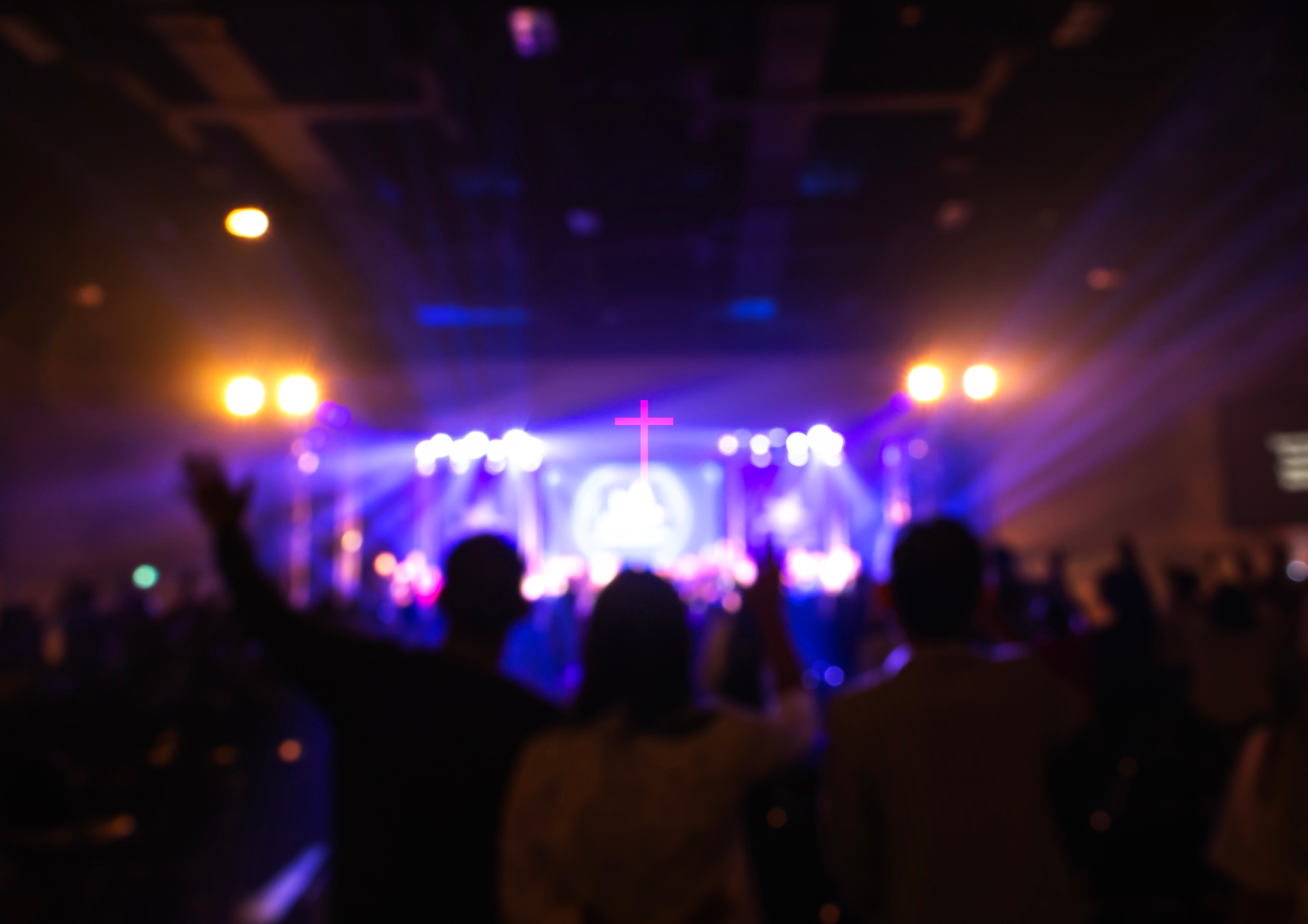 Silhouetted crowd at a concert with stage lights and a cross visible in the background