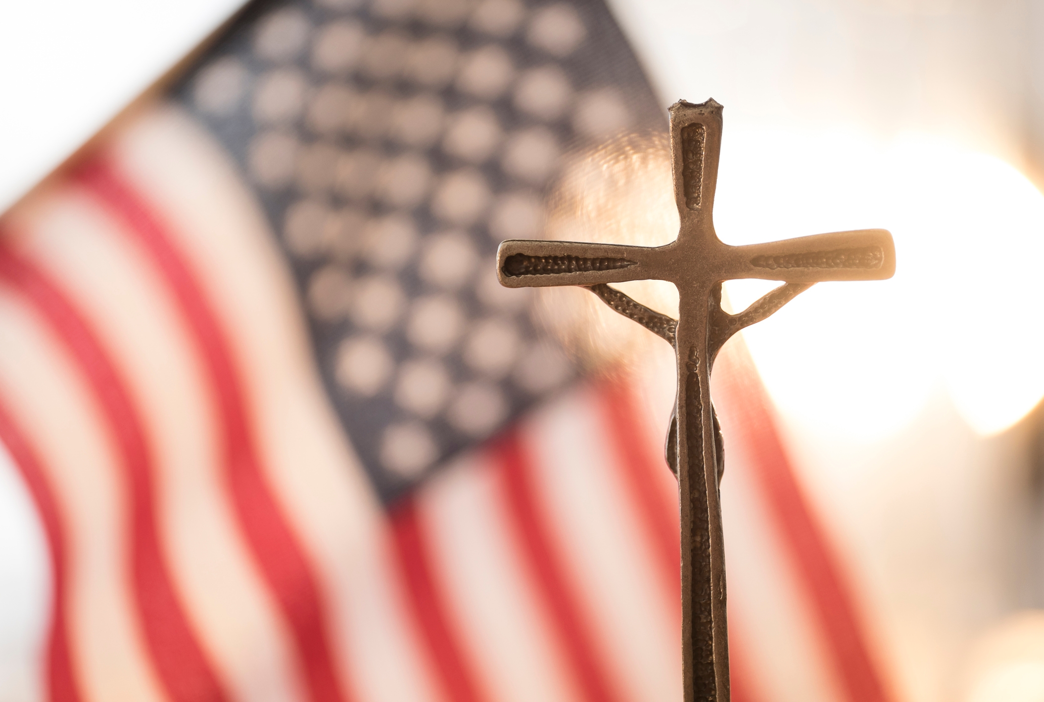 A cross in the foreground with the American flag blurred in the background