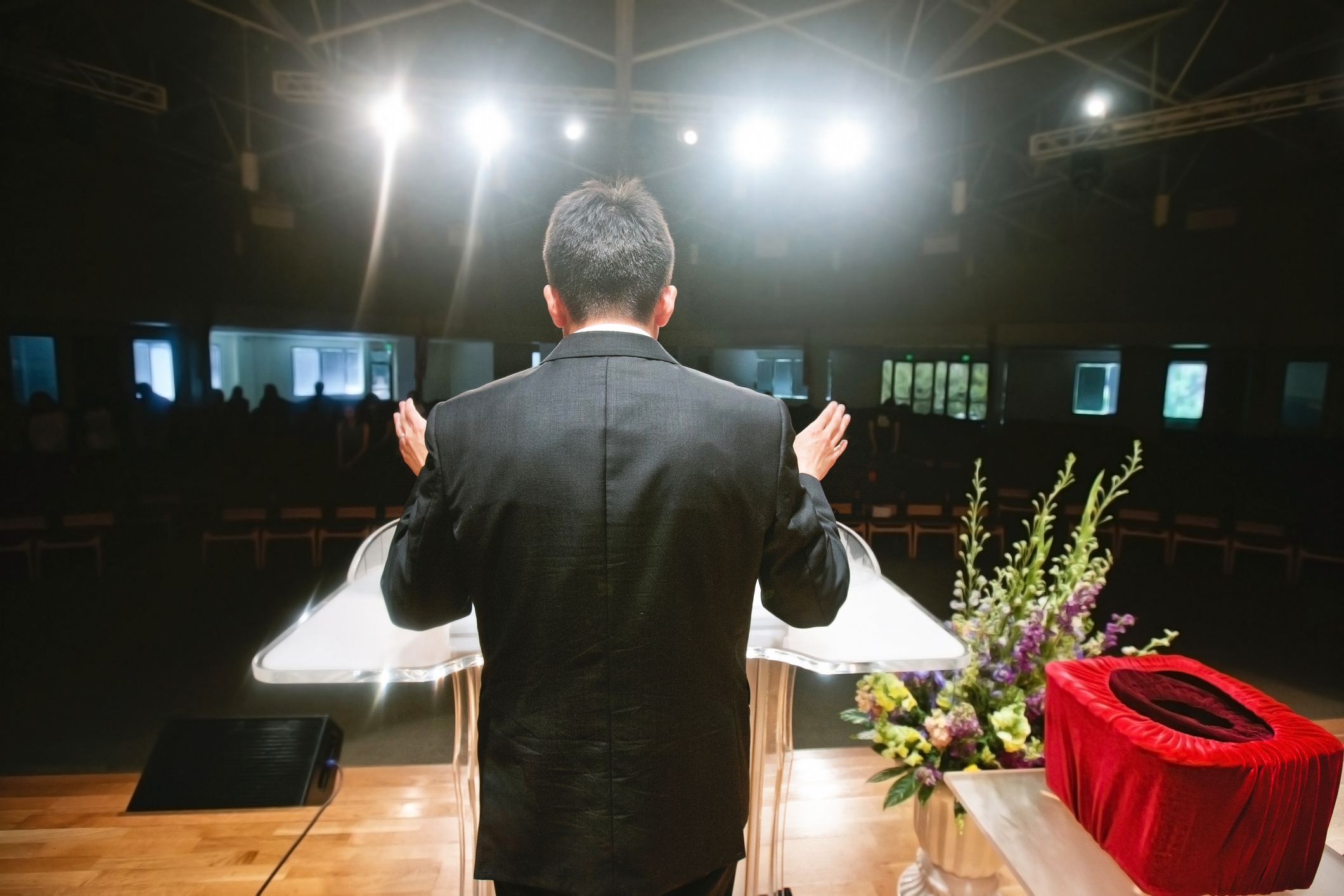 A man in a suit stands at a podium in a dimly lit auditorium, gesturing with hands raised, with an audience in silhouette