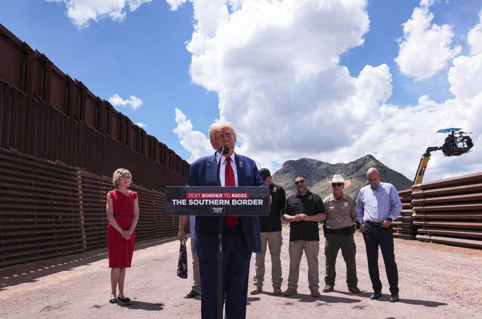 Group, including Trump in a suit speaking at a podium labeled "The Southern Border," stands near border wall with mountains in background