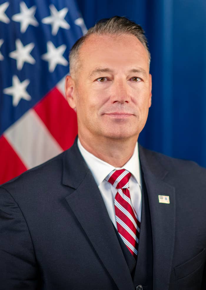 A person in a suit with a striped tie stands in front of a U.S. flag. Their expression is neutral