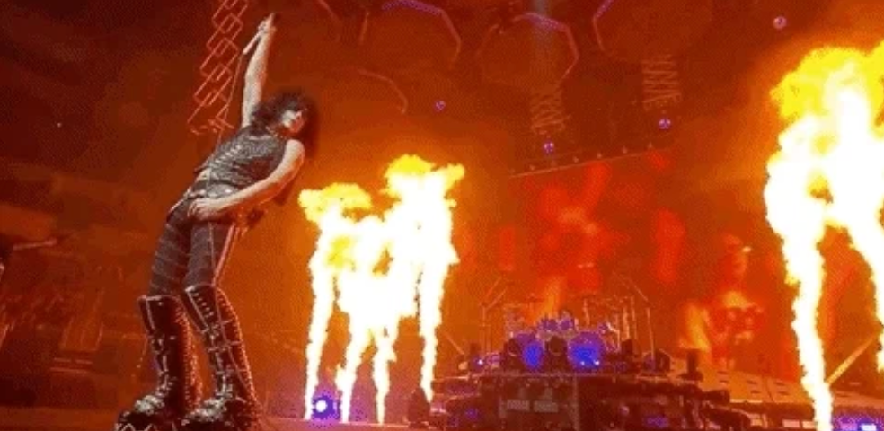 Performer in leather attire on stage, raising a fist amid pyrotechnics with a drum set in the background