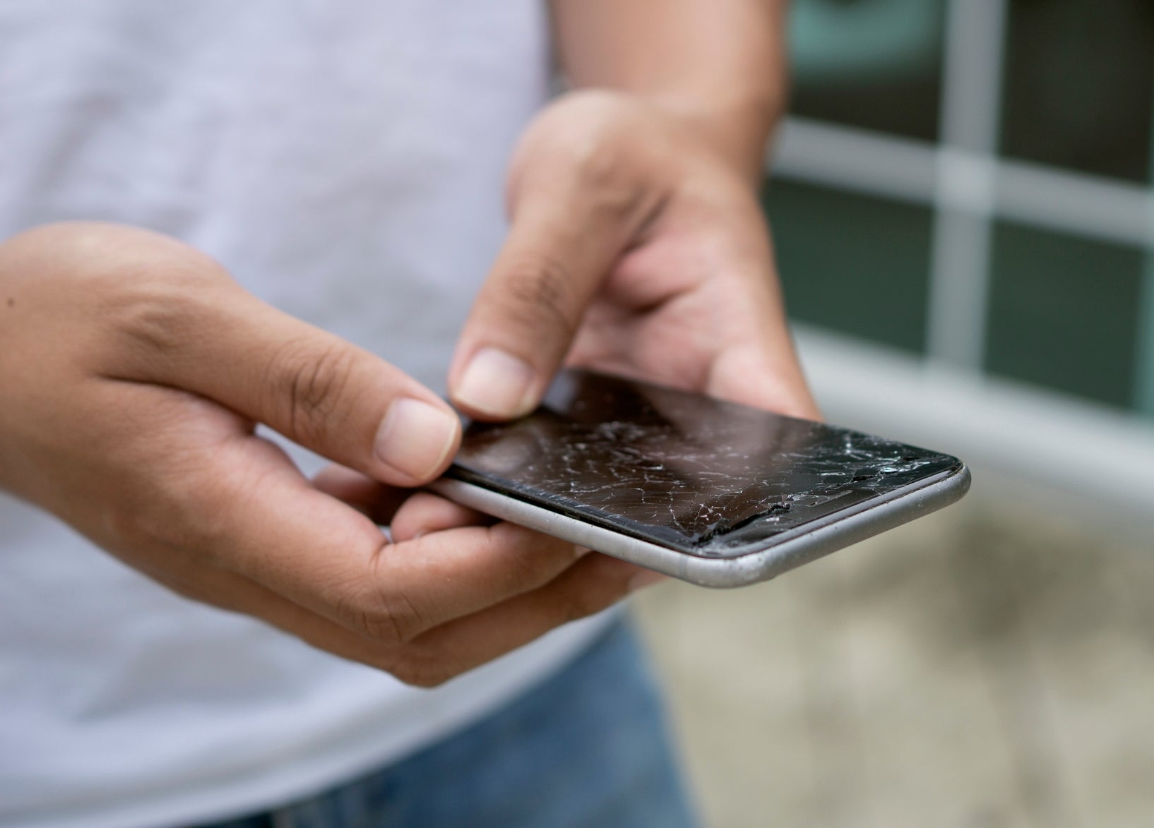 Person holding a smartphone with a cracked screen outdoors, suggesting issues related to repair costs or phone durability
