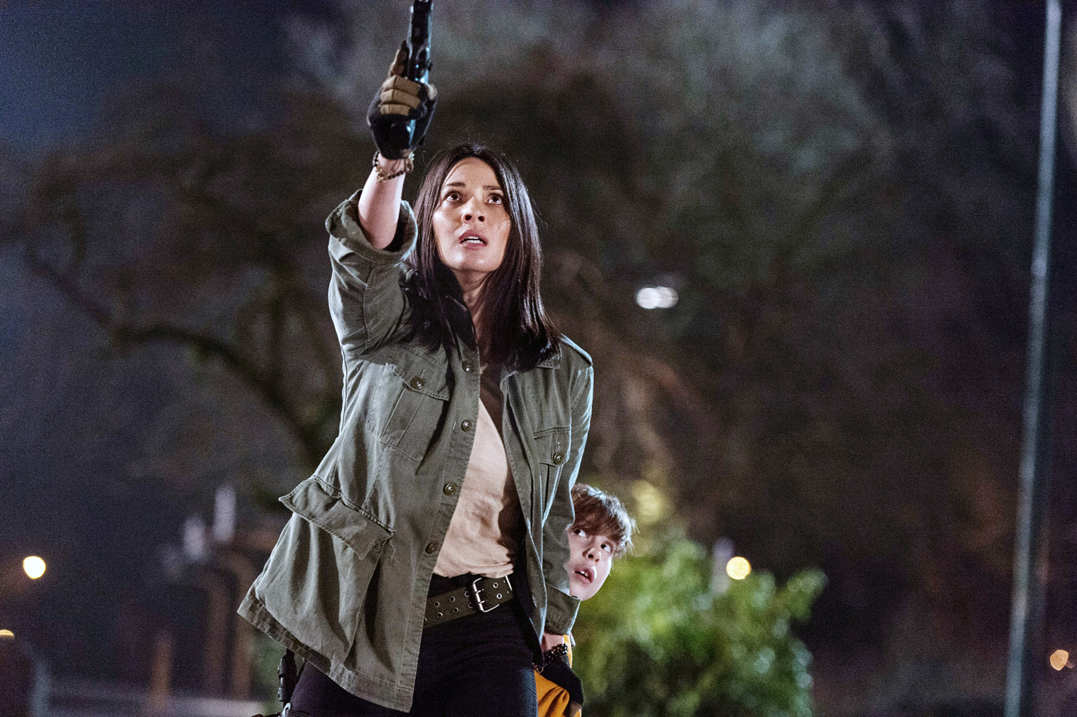 Person in a casual military-style jacket holds a gun up, with a child behind them, both appearing focused in a night-time scene