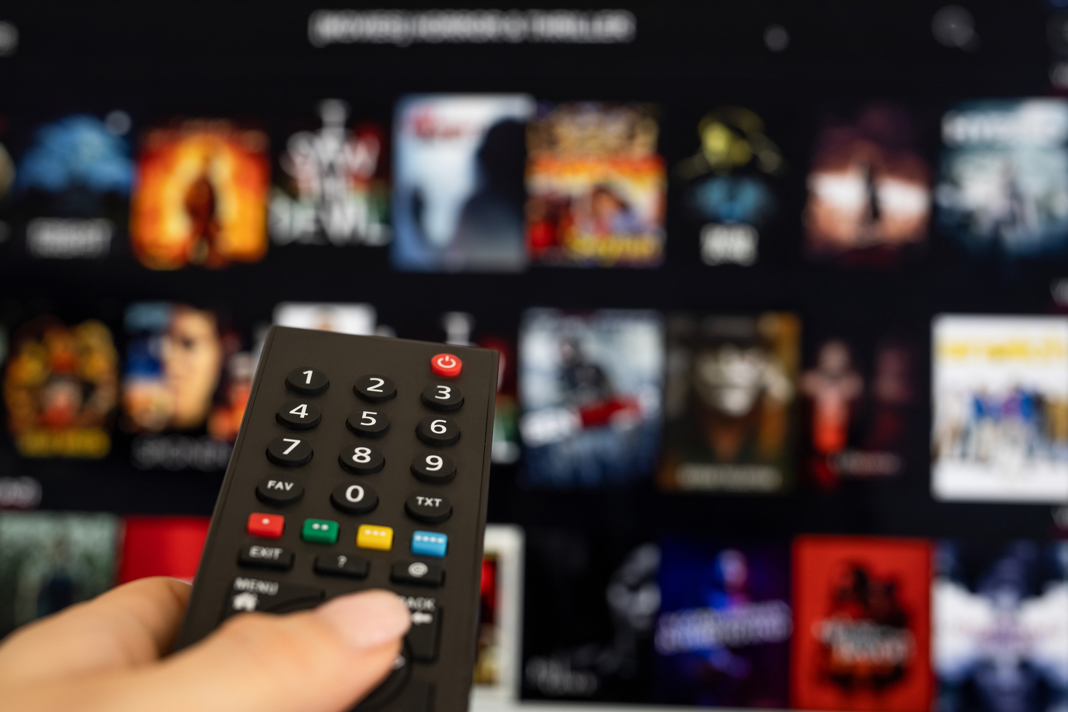A hand holding a TV remote points at a streaming service menu displaying various movie and show thumbnails on a screen