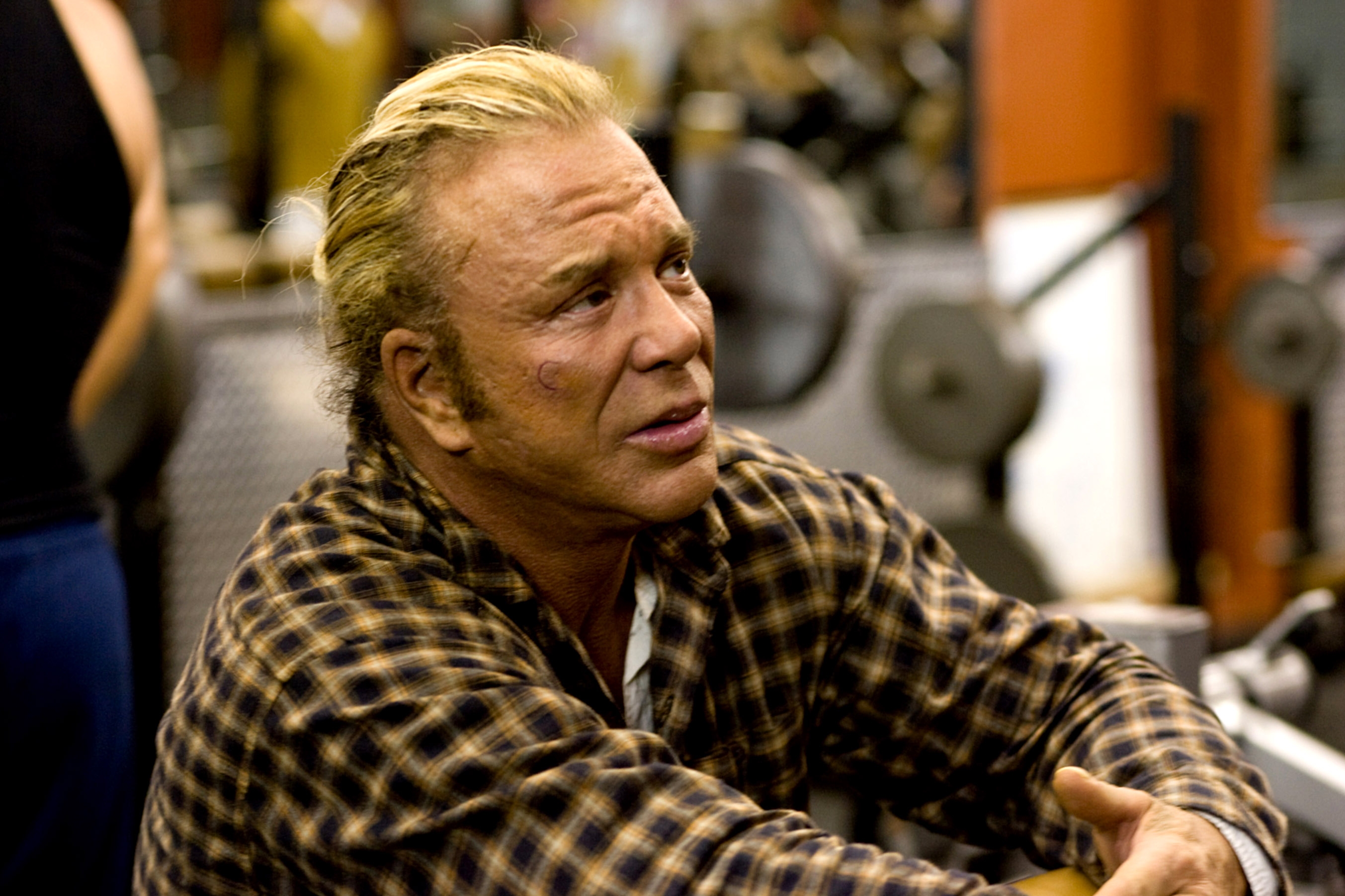 Person in a checkered shirt sitting in a gym setting, appearing thoughtful with workout equipment in the background