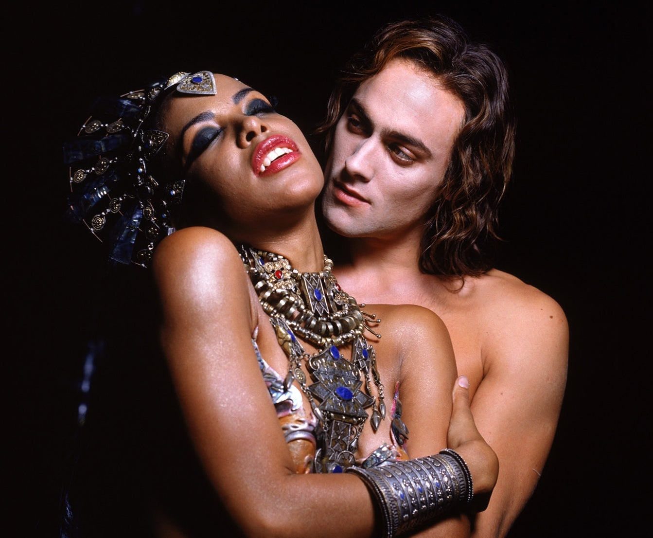 A person in elaborate jewelry, embraced by a shirtless individual, both with dramatic poses