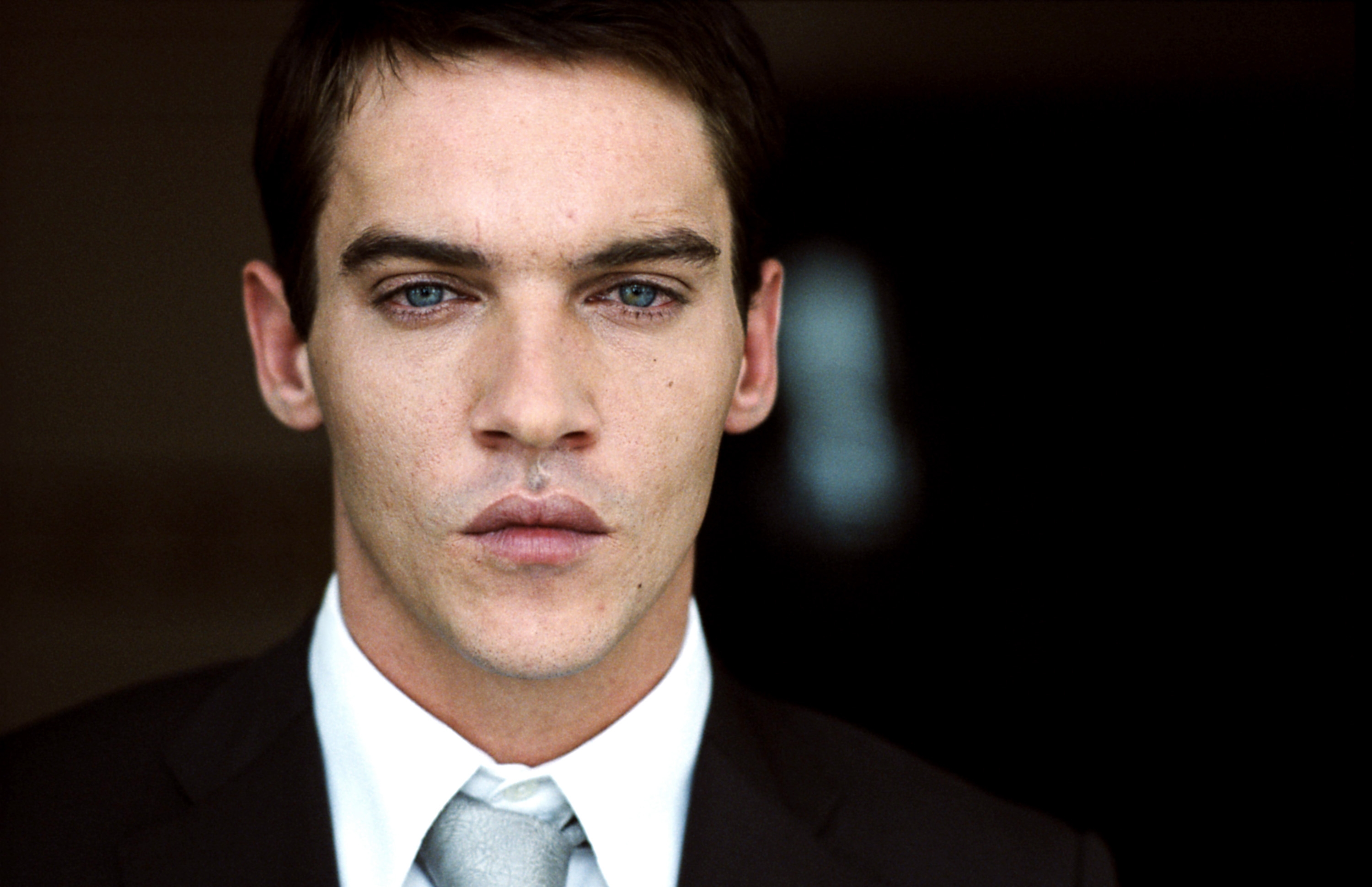 A person in a formal suit with a tie, standing against a plain background