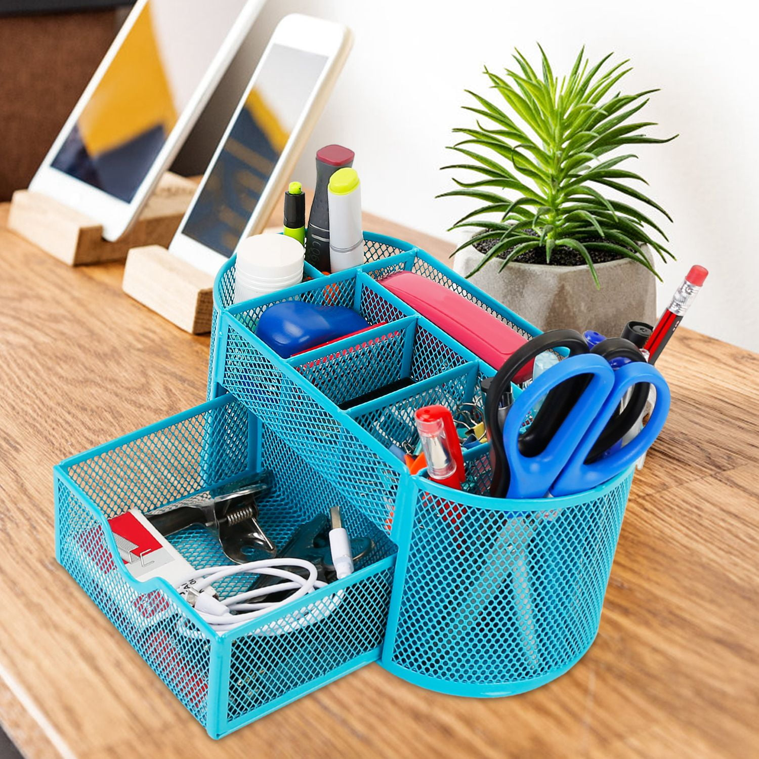 Desktop organizer with stationery items, including scissors, pens, and sticky notes, sits on a wooden desk near a small plant and tablets