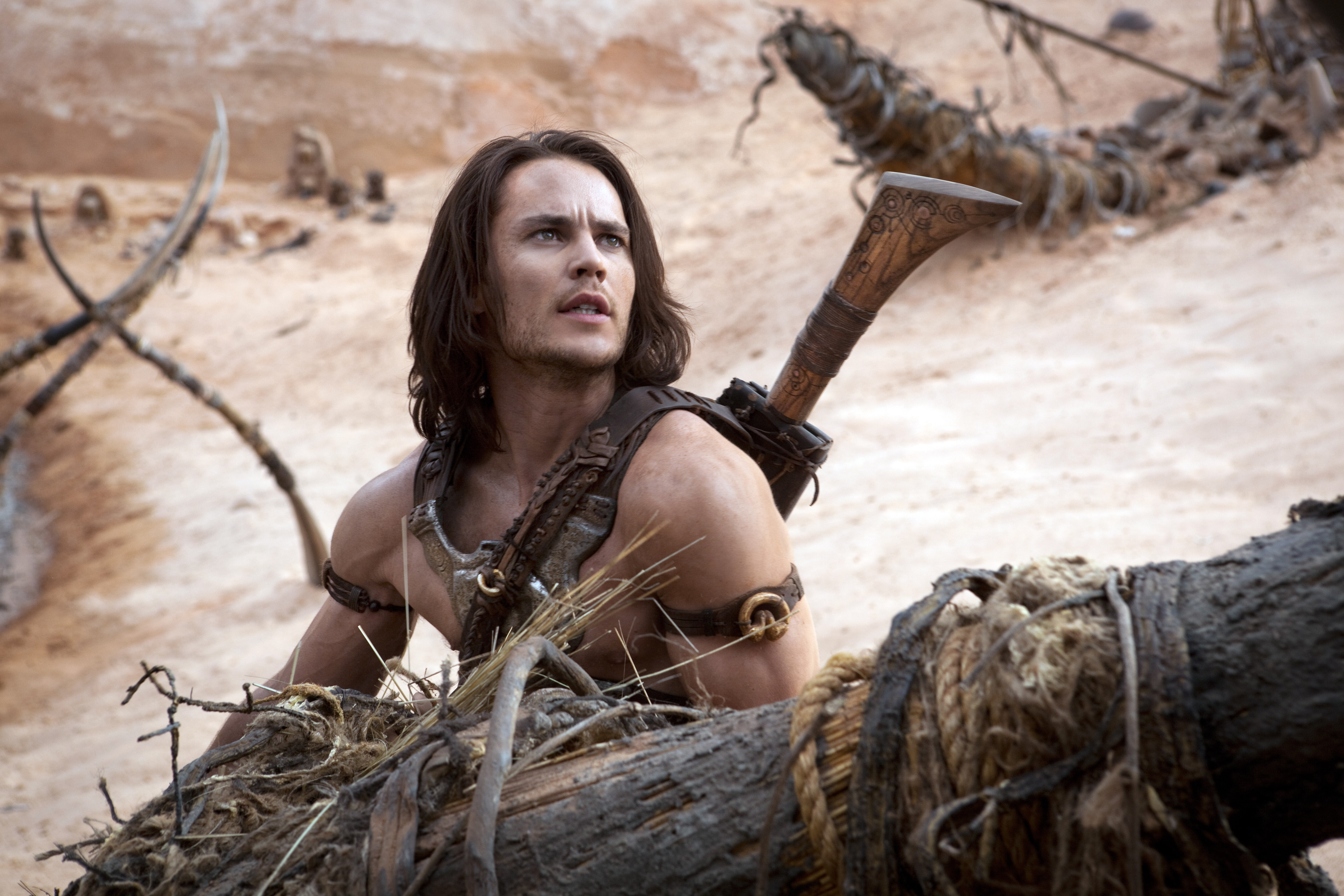 A man with long hair and a rugged look, dressed in primitive warrior attire, is perched on a large branch in a barren landscape