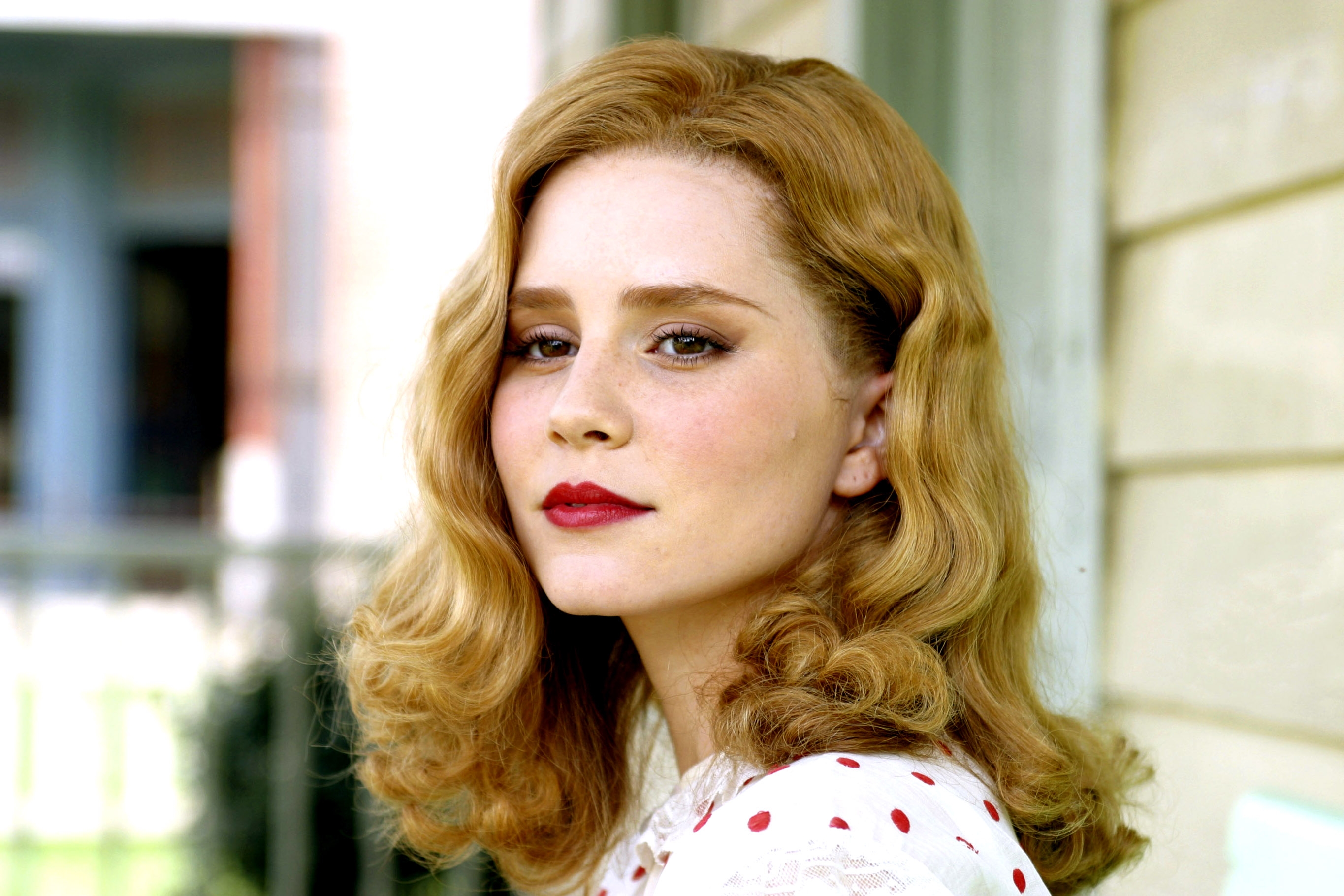 Person with wavy hair and bright lipstick, wearing a polka-dotted top, outside a building