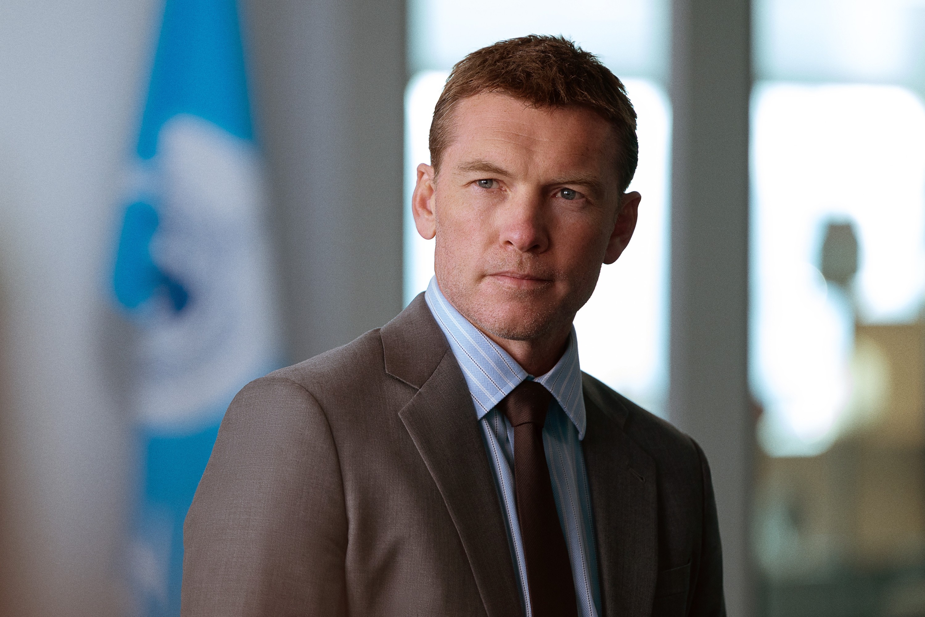 Person in a suit looks slightly to the side, standing indoors with a blurred flag in the background