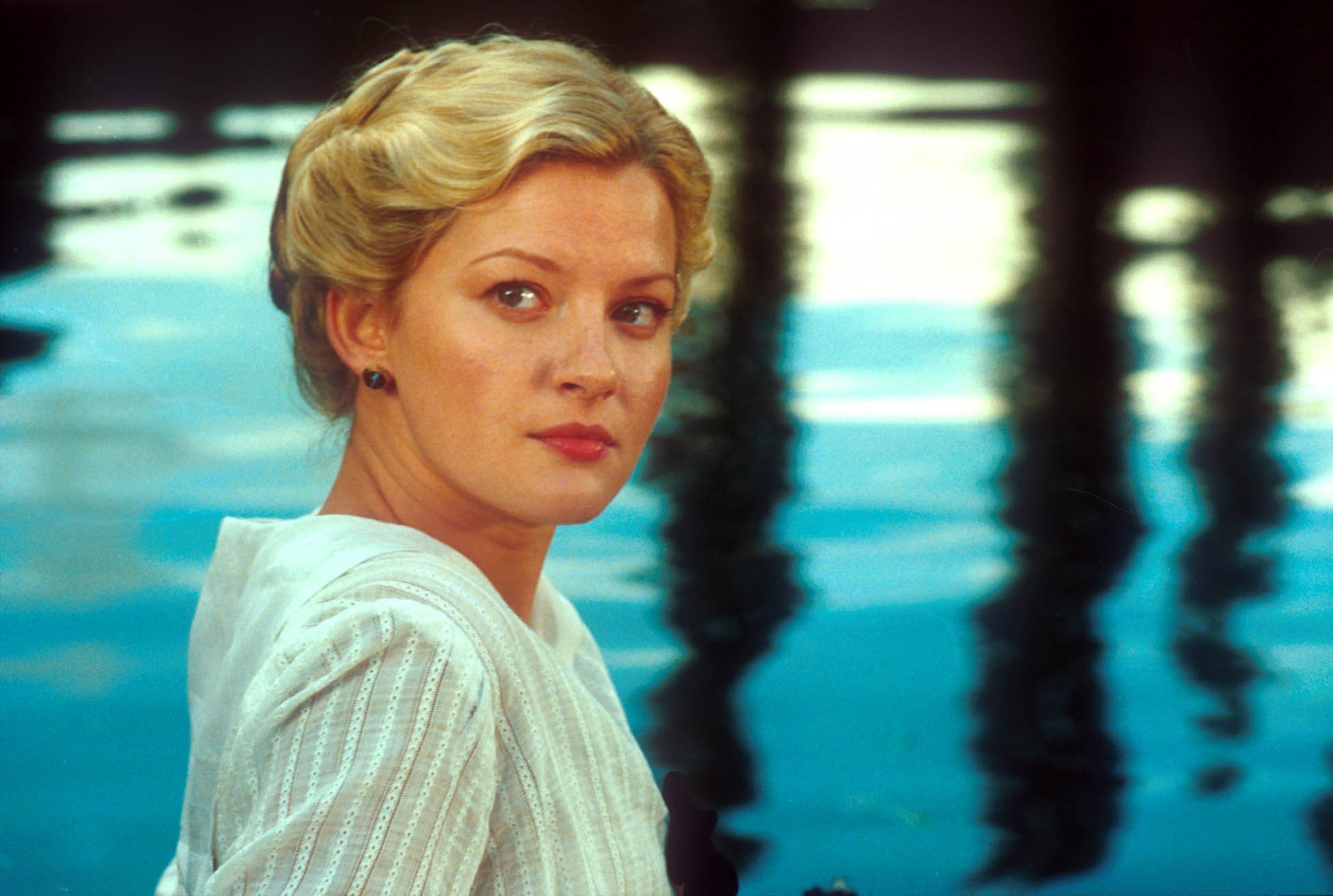 Person by a pool, with neatly styled hair, wearing a light-colored outfit with subtle stripes, looking over their shoulder