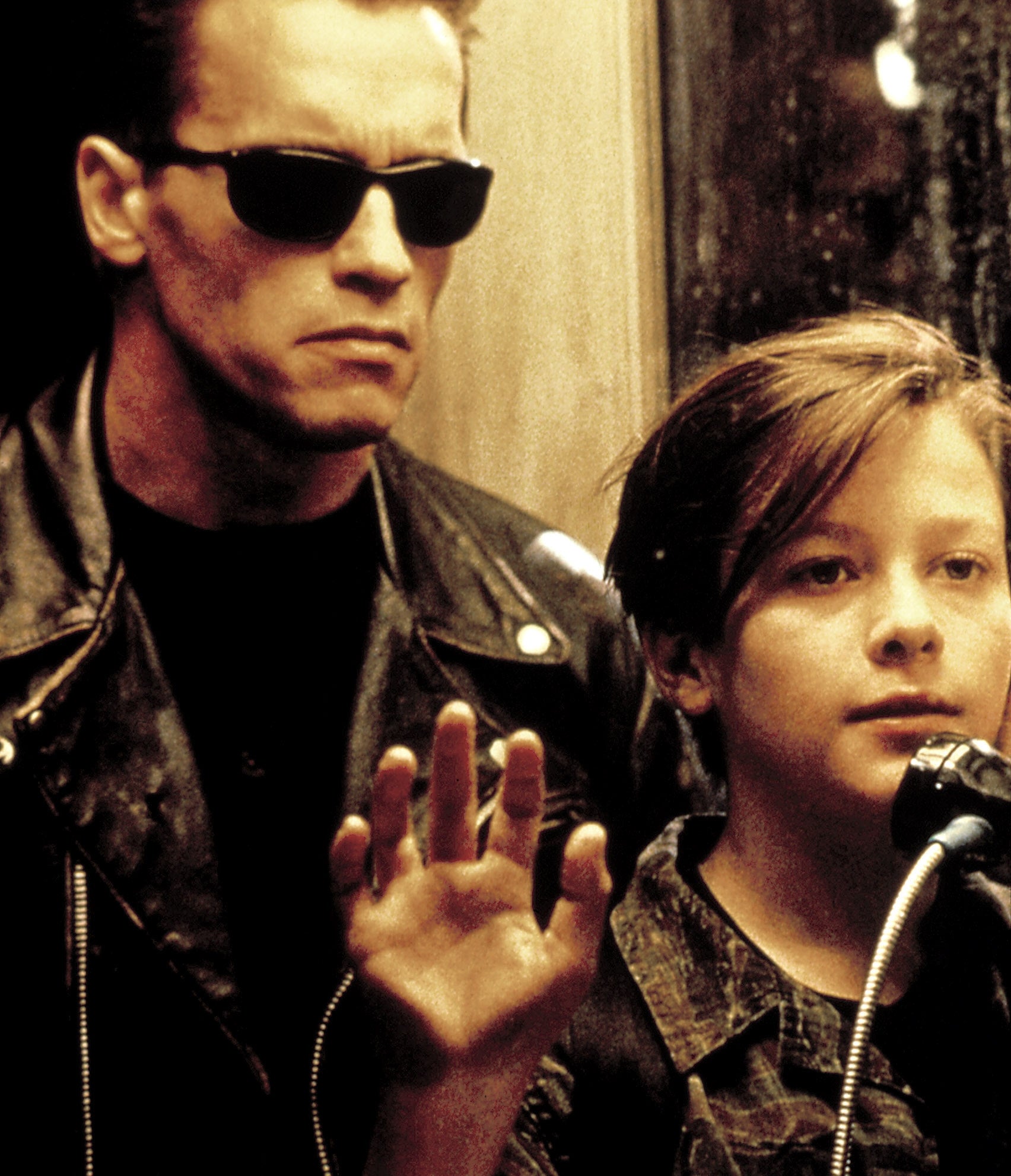 A man in a leather jacket and sunglasses stands beside a boy using a payphone, in a scene from an action movie
