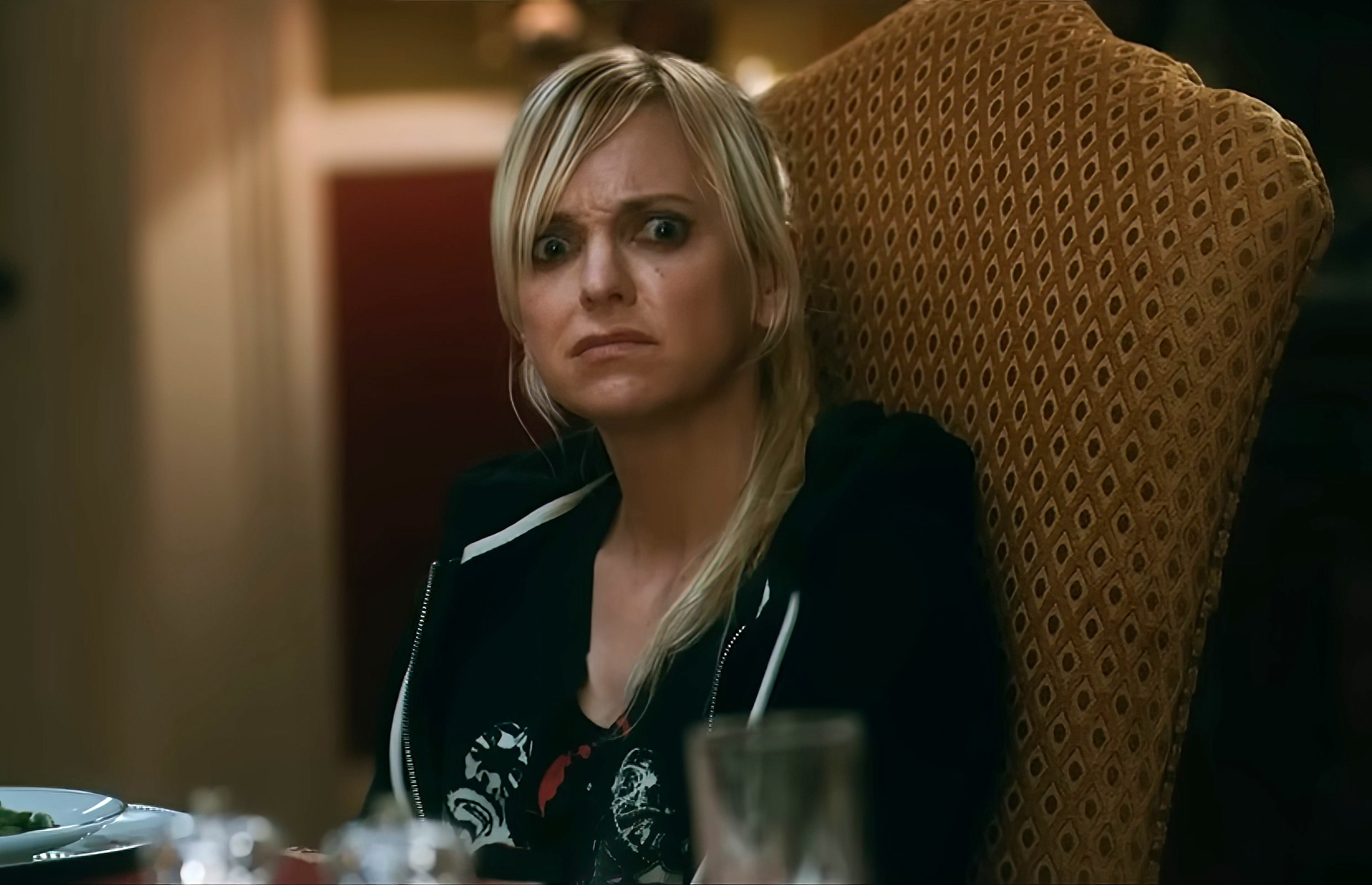 Person seated at a dining table, looking shocked and concerned, wearing a graphic t-shirt and zip-up jacket