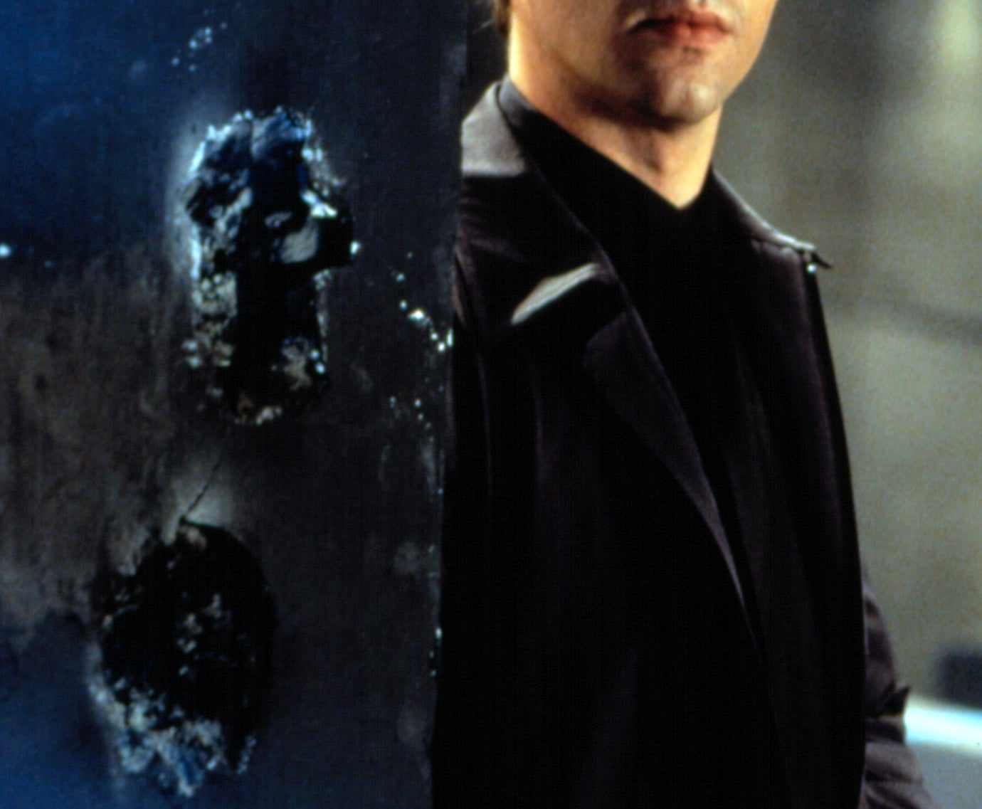 A person in a dark outfit stands partially hidden behind a wall with bullet holes, holding a small object, looking alert