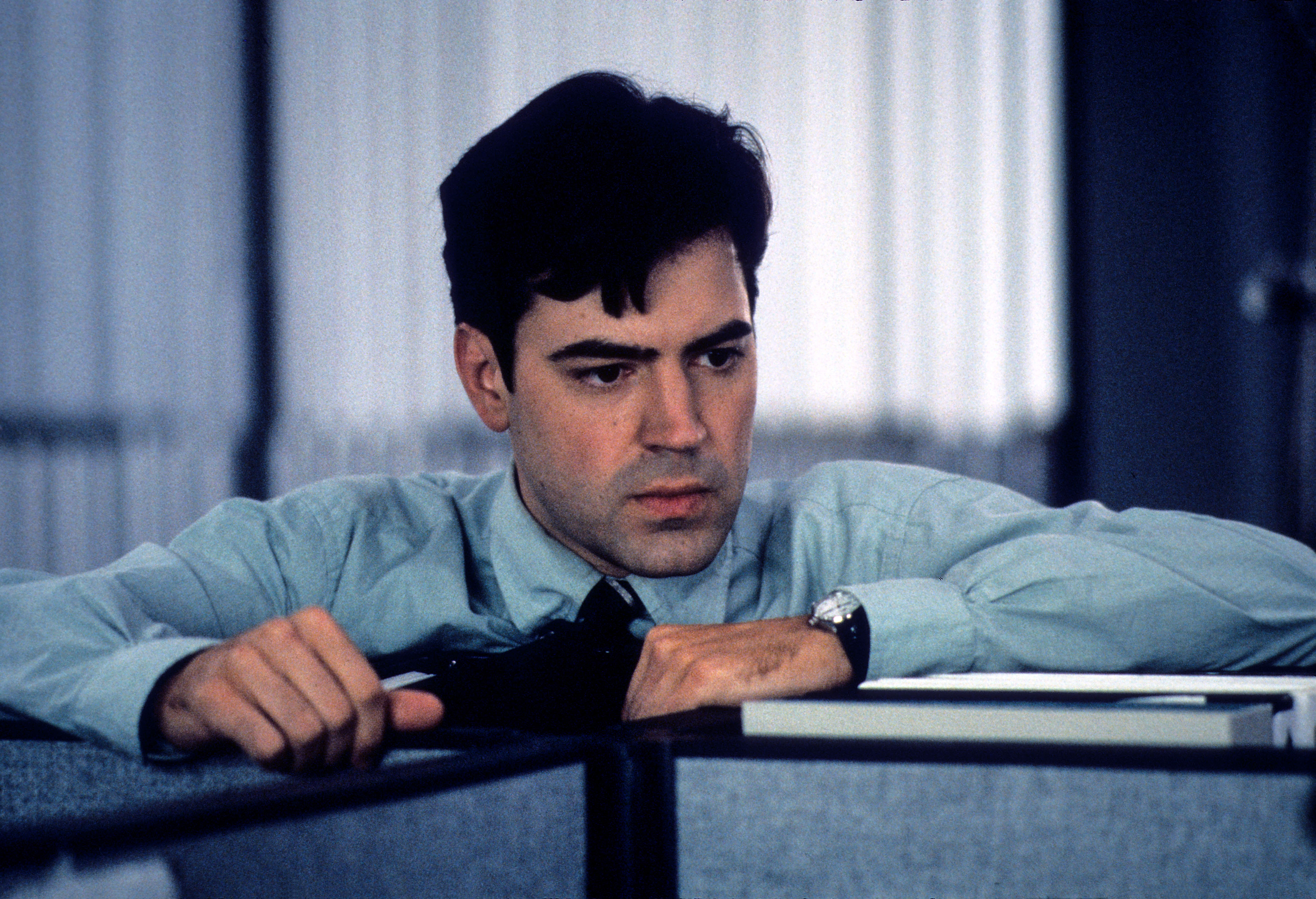 Person in an office setting, looking contemplative over a cubicle wall, wearing a dress shirt and tie