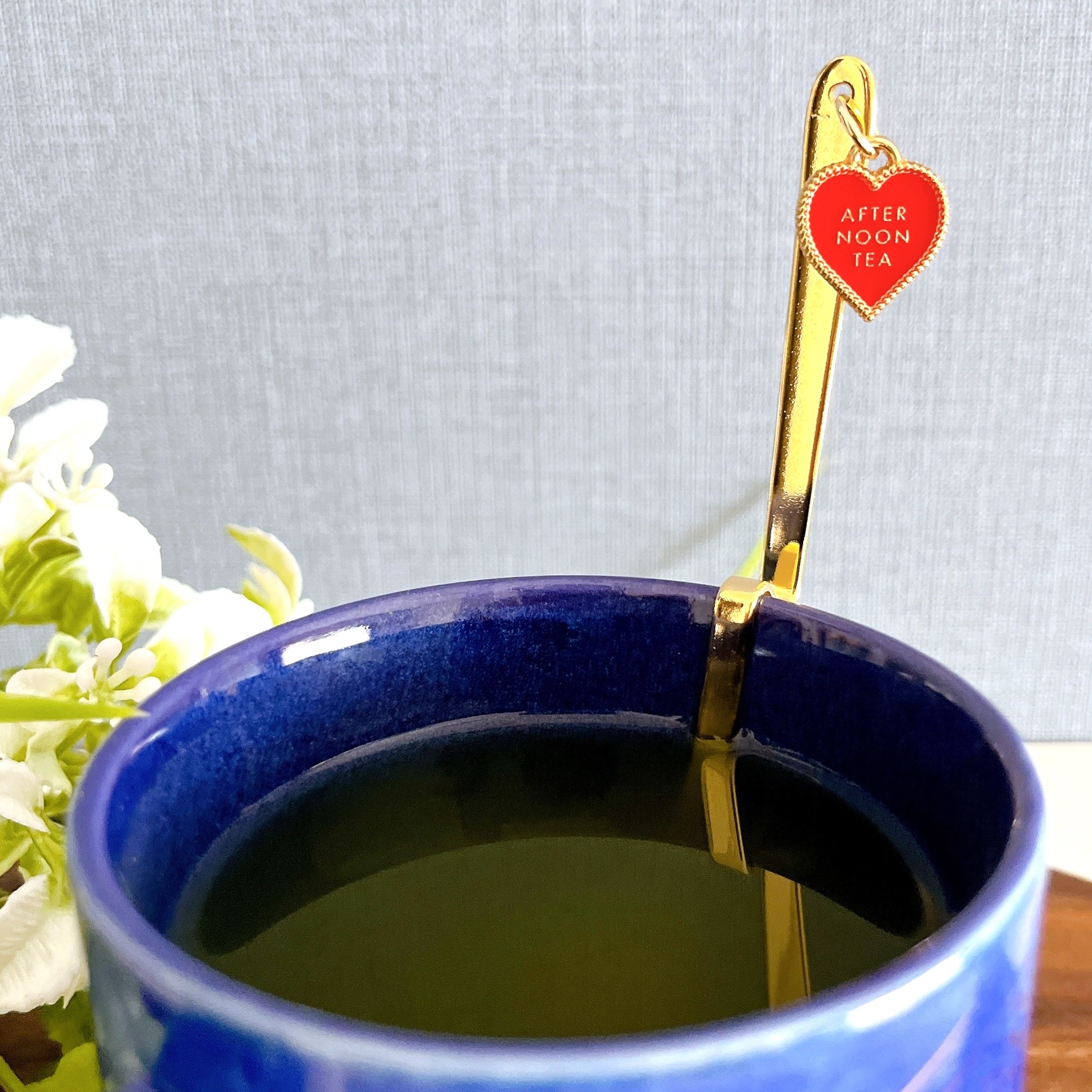 青いカップに入ったお茶と、「AFTERNOON TEA」と書かれた赤いハートがついた金色のスプーンが見える。白い花も添えられている。