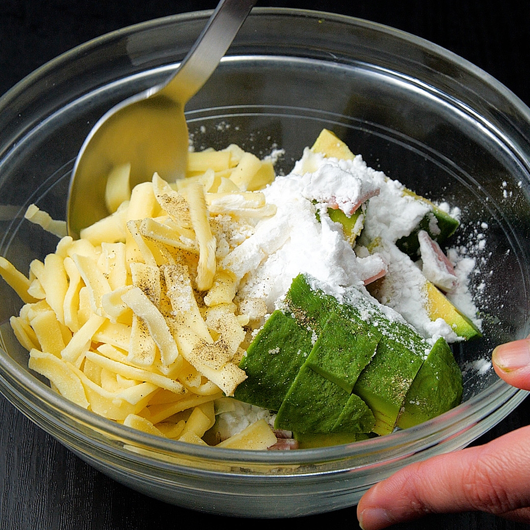 調理中のガラスボウルに麺、生アボカド、粉末調味料が入った様子。手がボウルを支えている。