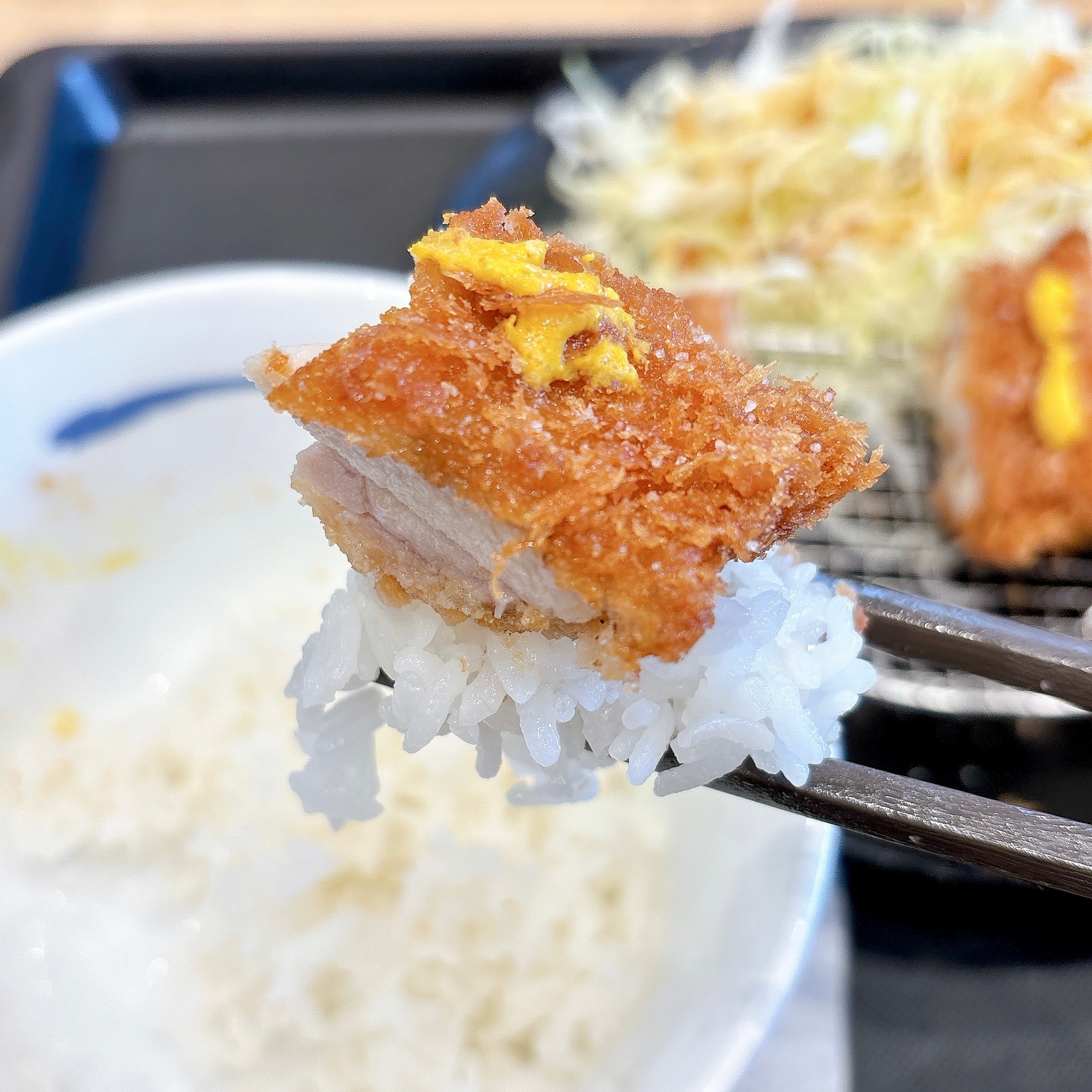 とんかつの一片がご飯とともに箸で持ち上げられている。背景にキャベツのつけあわせが見える。