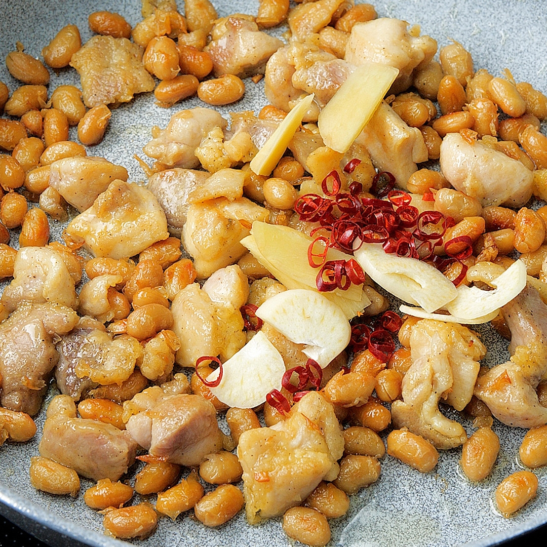 豆と鶏肉の炒め物が皿に盛られている。スライスされたニンニクと唐辛子がトッピングされている。