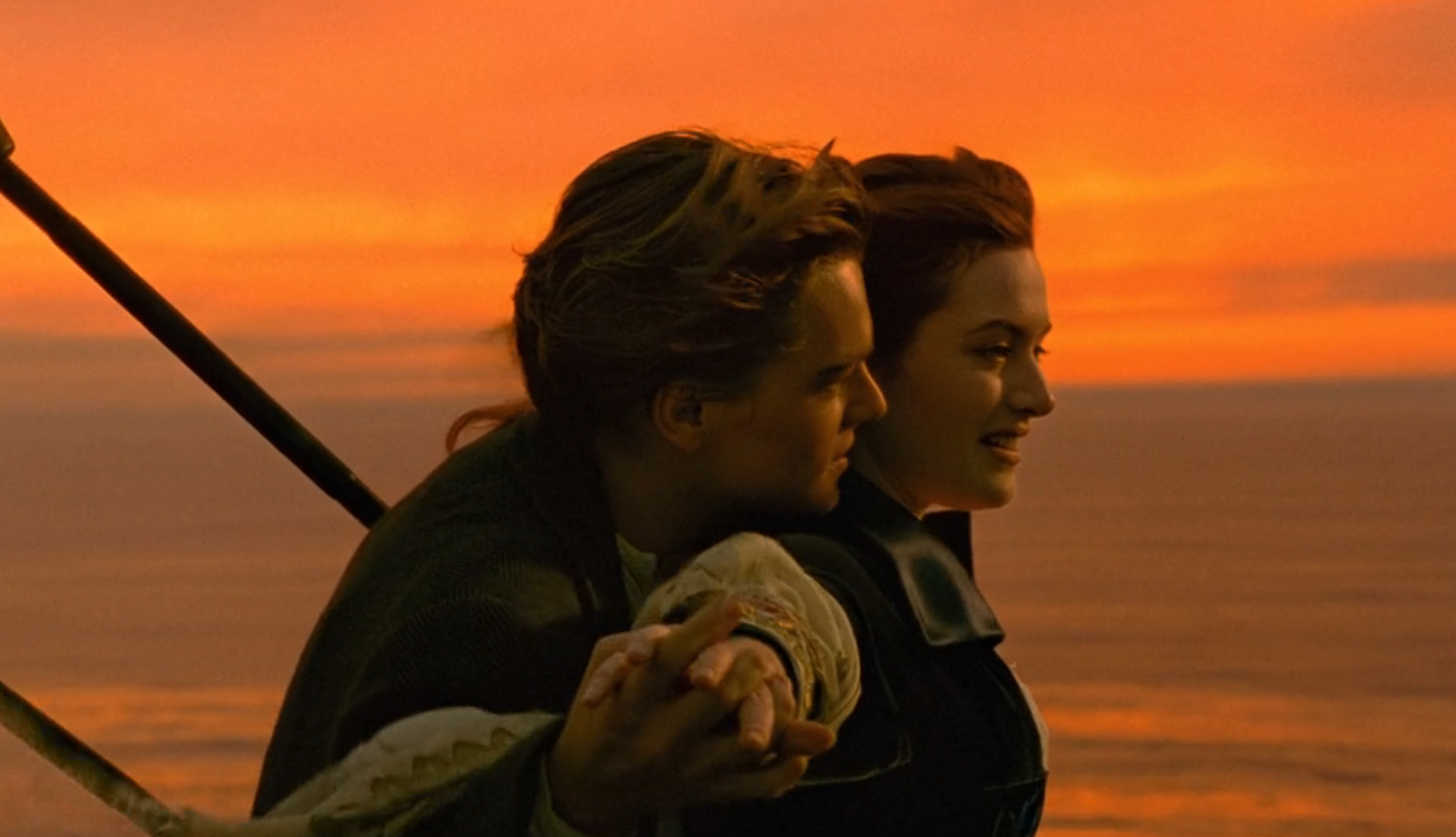 A couple stands at the ship's bow, arms outstretched, embracing as they enjoy a scenic sunset over the ocean