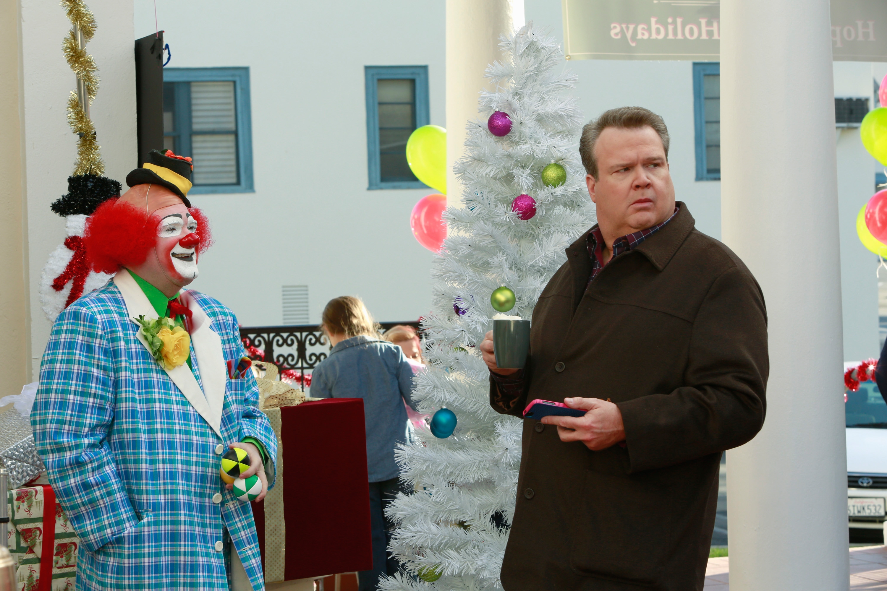 A man in a coat looks surprised next to a clown in a plaid suit during a holiday event with a white Christmas tree and colorful balloons