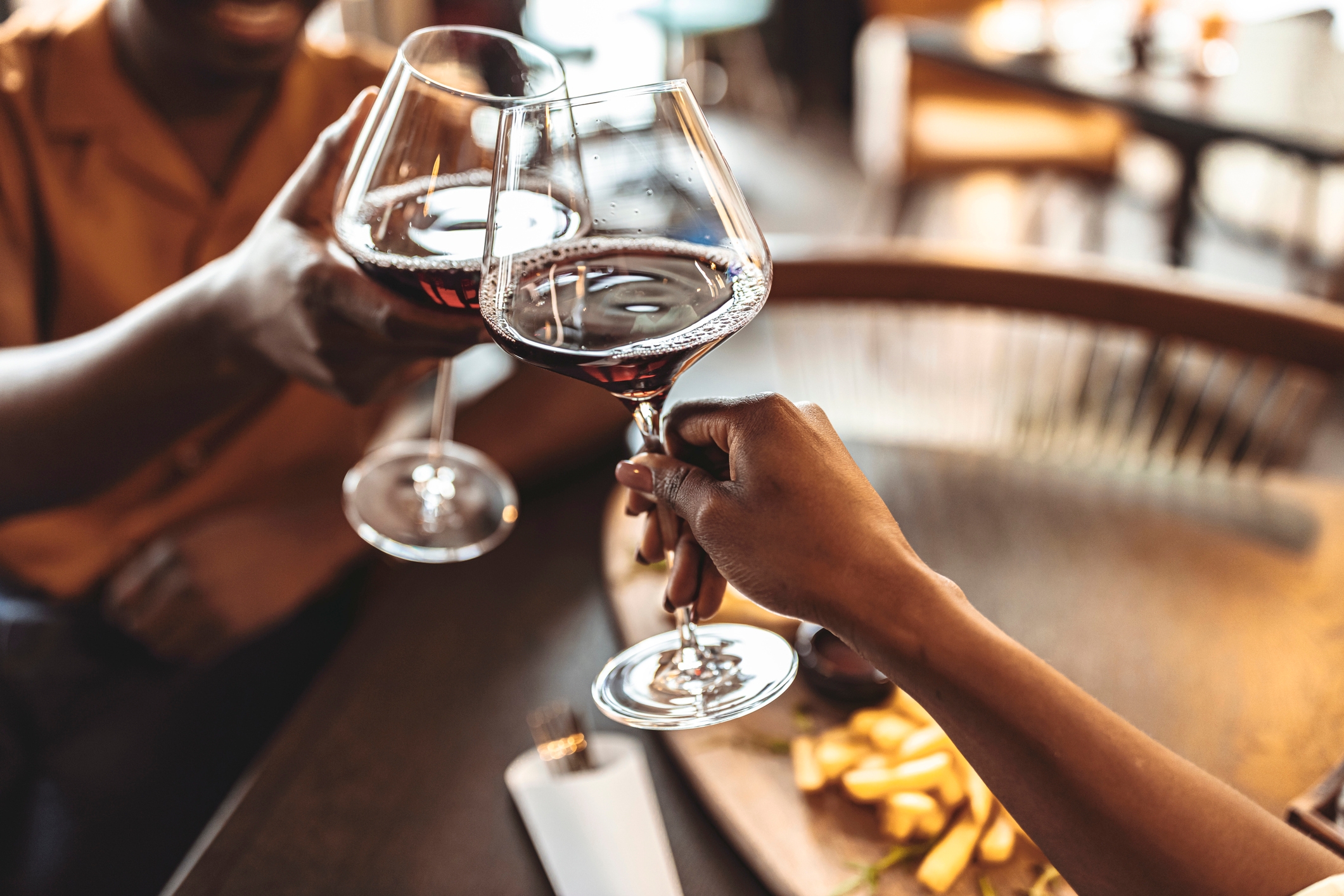 Two people clinking wine glasses over a table, with fries in the background, suggesting a romantic dining setting