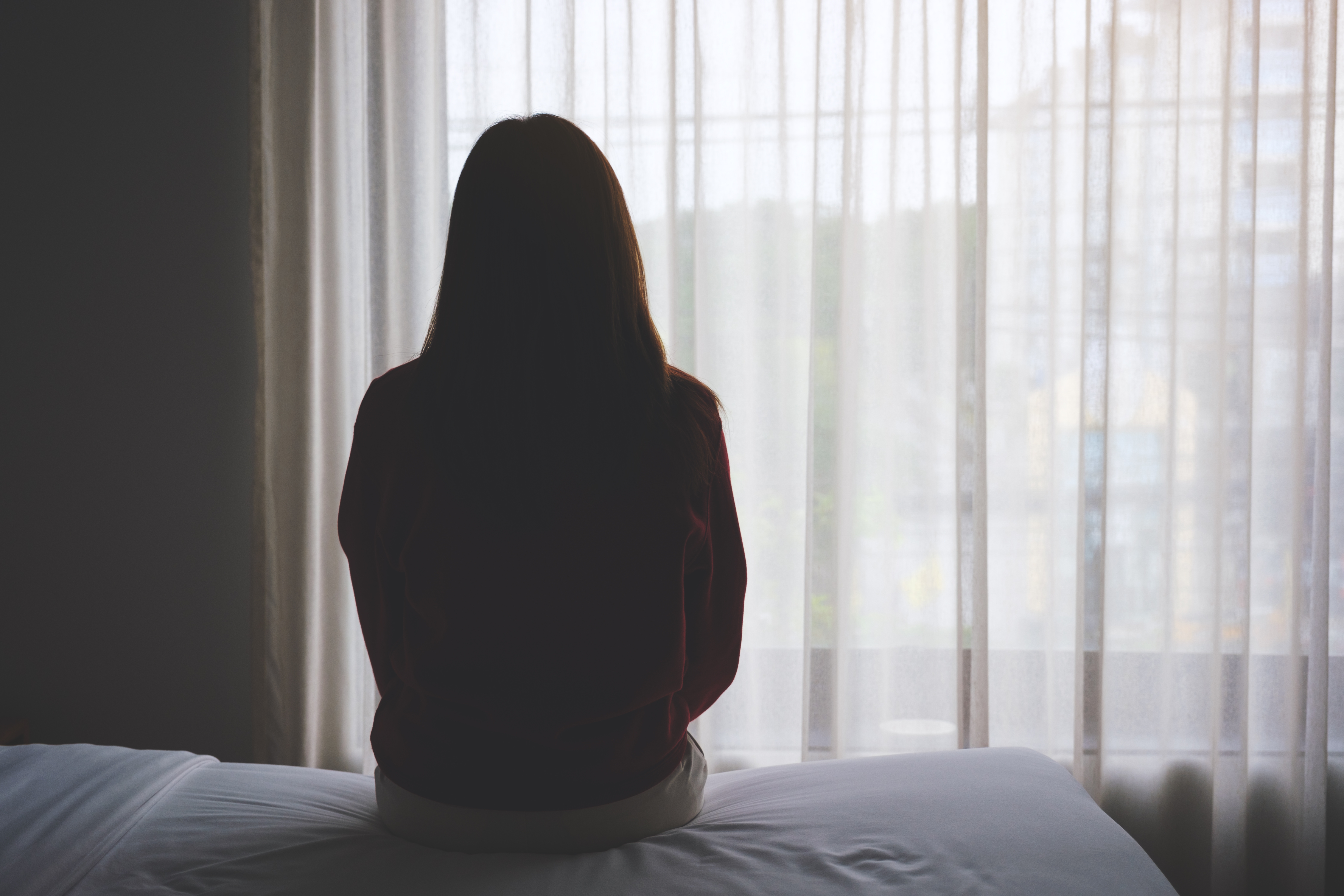 Silhouette of a person sitting on a bed, facing a window with sheer curtains.