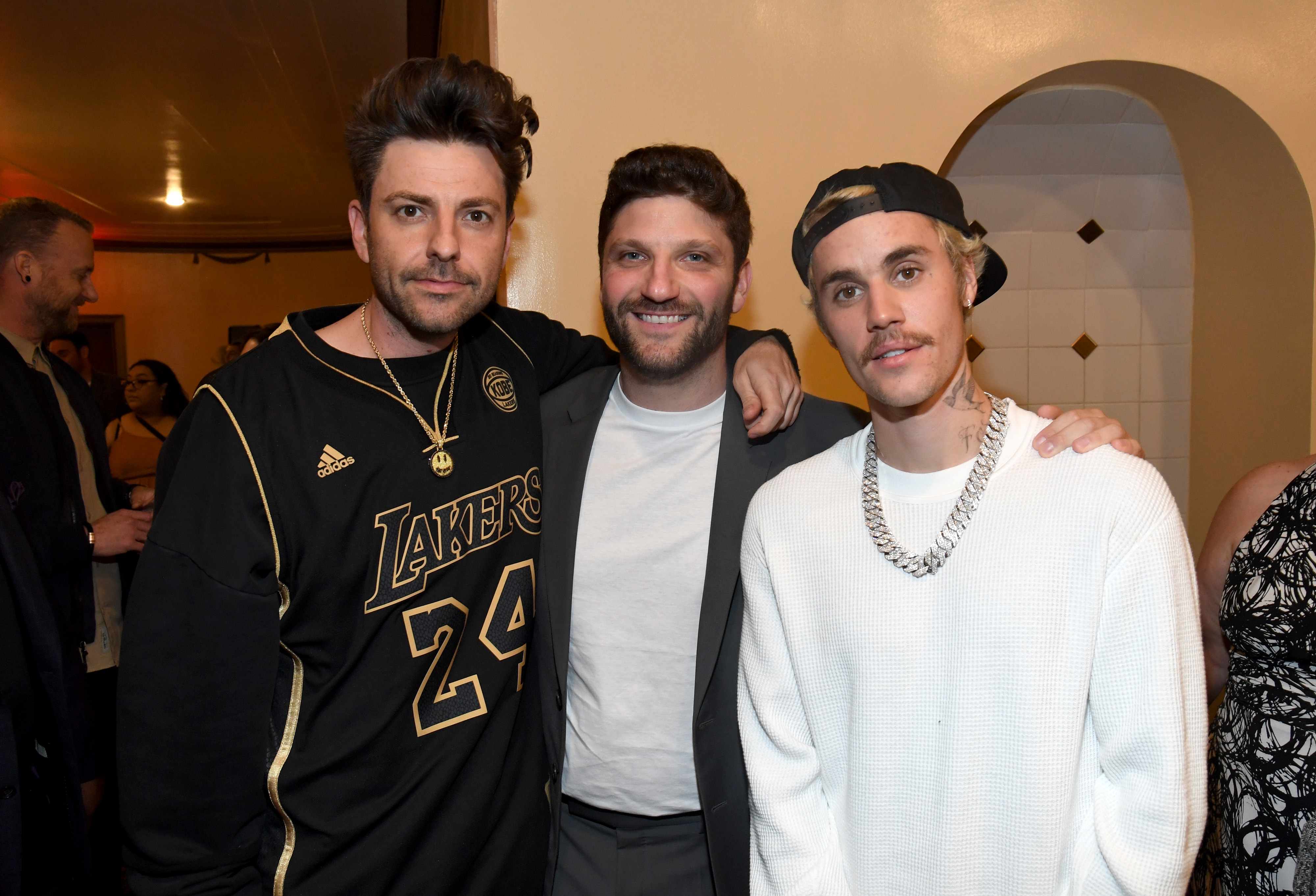 Three people pose together at an event. One wears a sports jersey, another is in a suit, and the third wears a casual outfit with a baseball cap