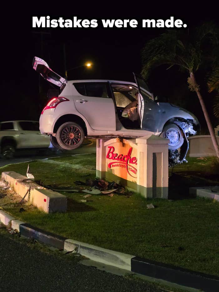 Car balanced precariously on roadside sign at night; it appears to have driven up and over a curb, coming to rest in a dramatic position