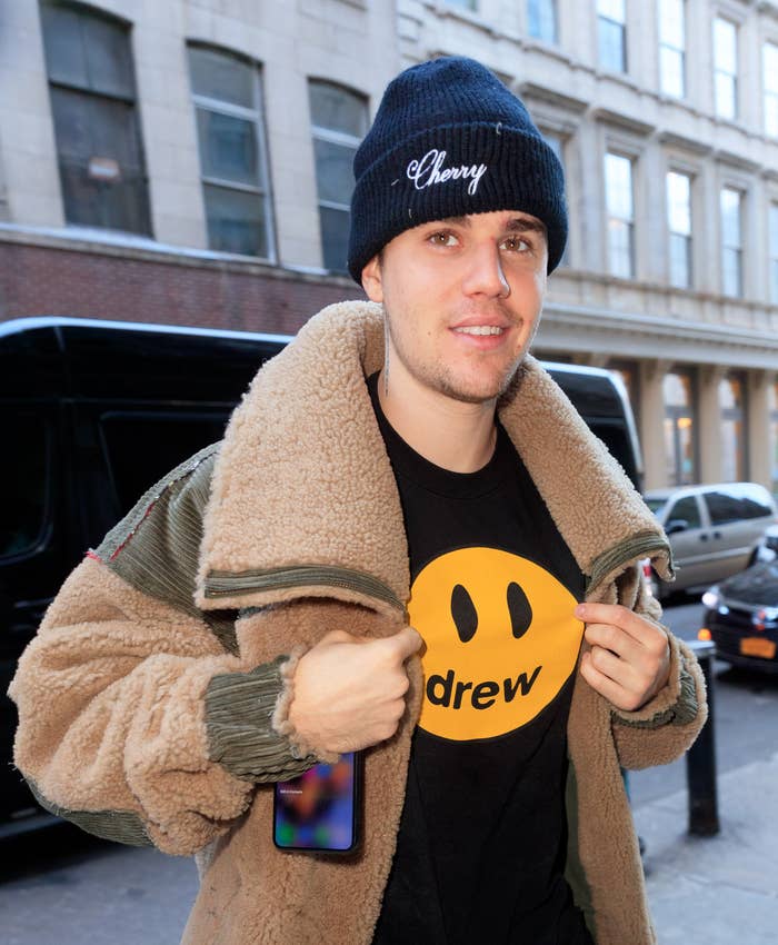 Person wearing a shearling jacket over a black t-shirt with a smiley face, beanie with text, casually walking in an urban setting