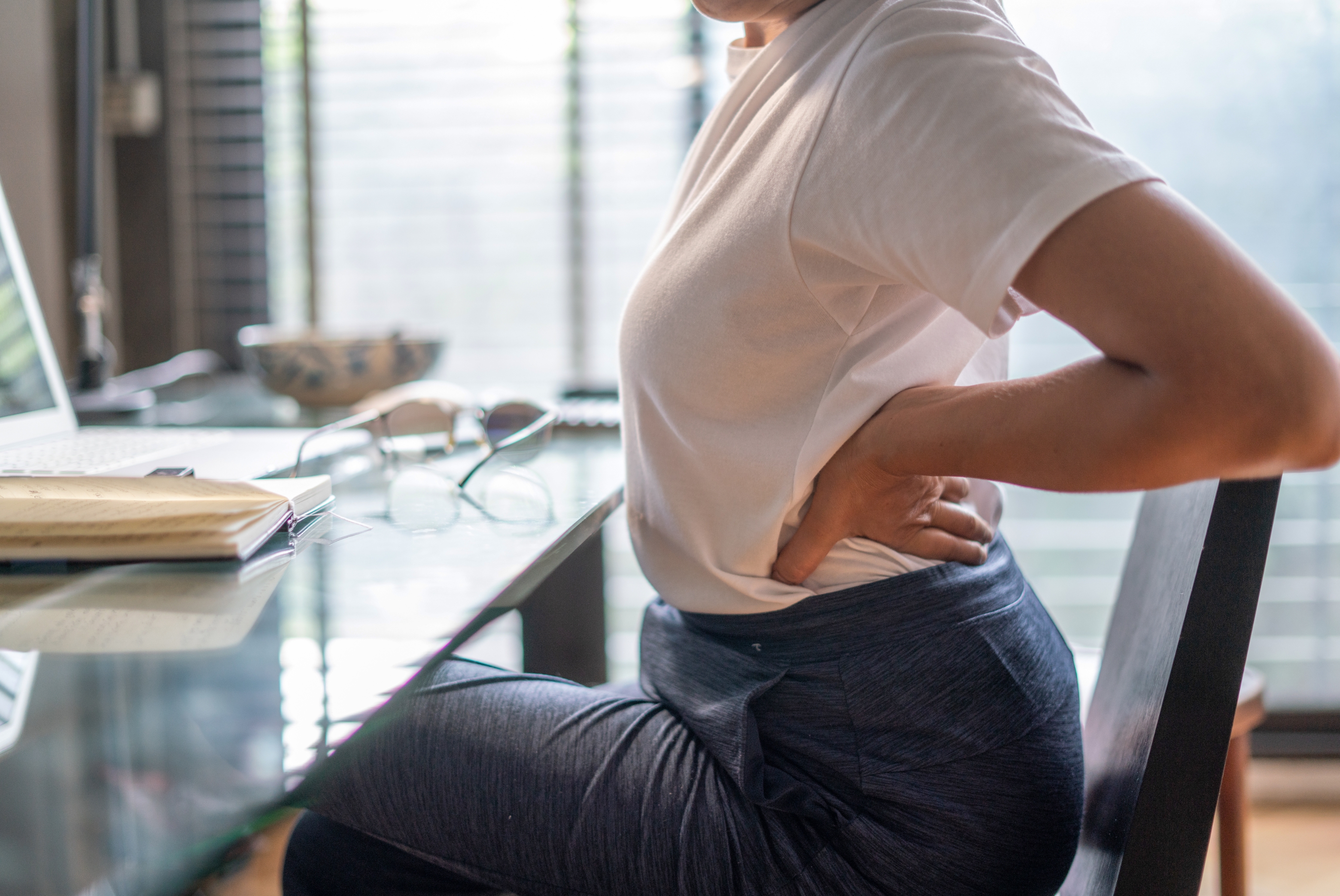Person seated at a desk, holding their back in discomfort, with a laptop, notebook, and glasses nearby.
