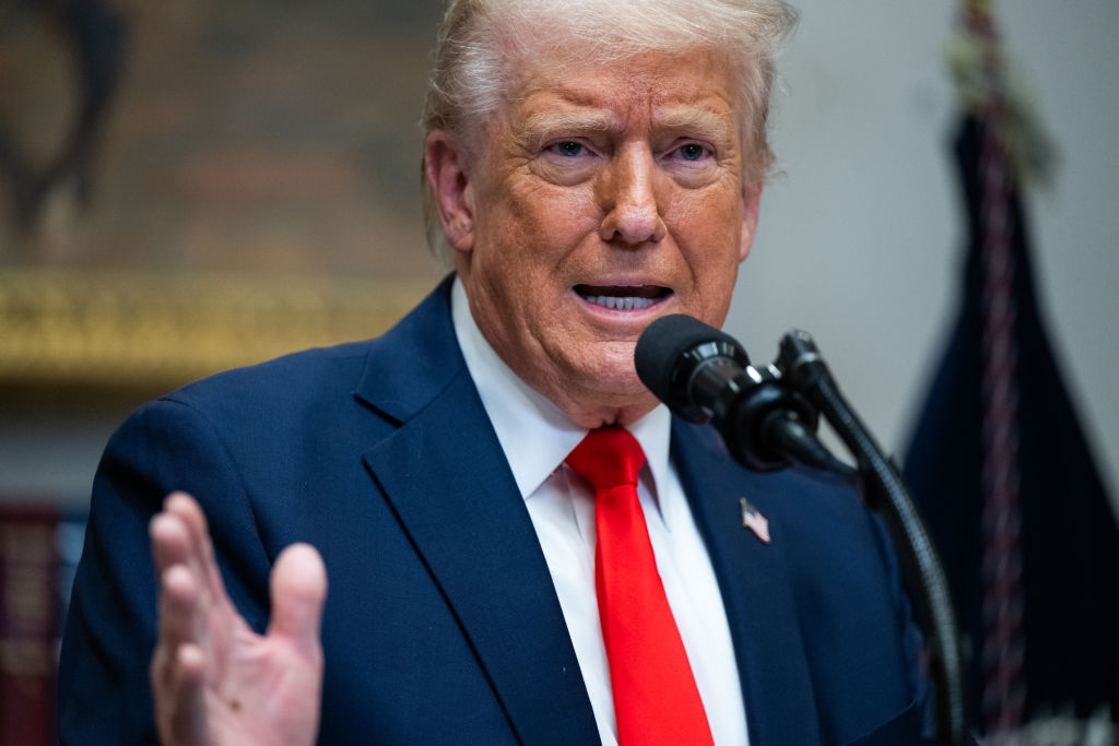 A person in a suit and red tie speaks at a microphone during a formal event