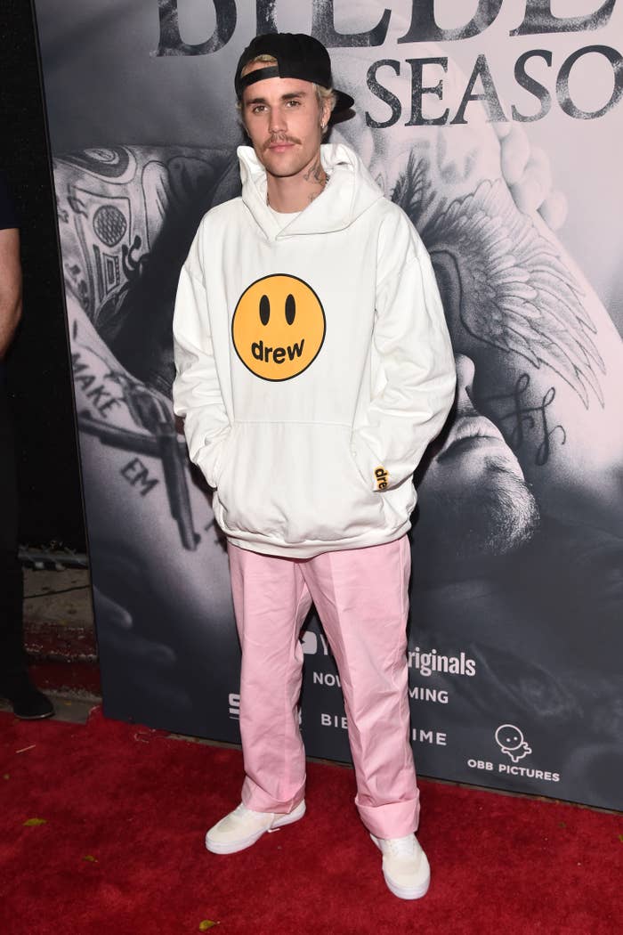 Person in casual attire with a white hoodie featuring a smiley face and pink pants, standing on a red carpet