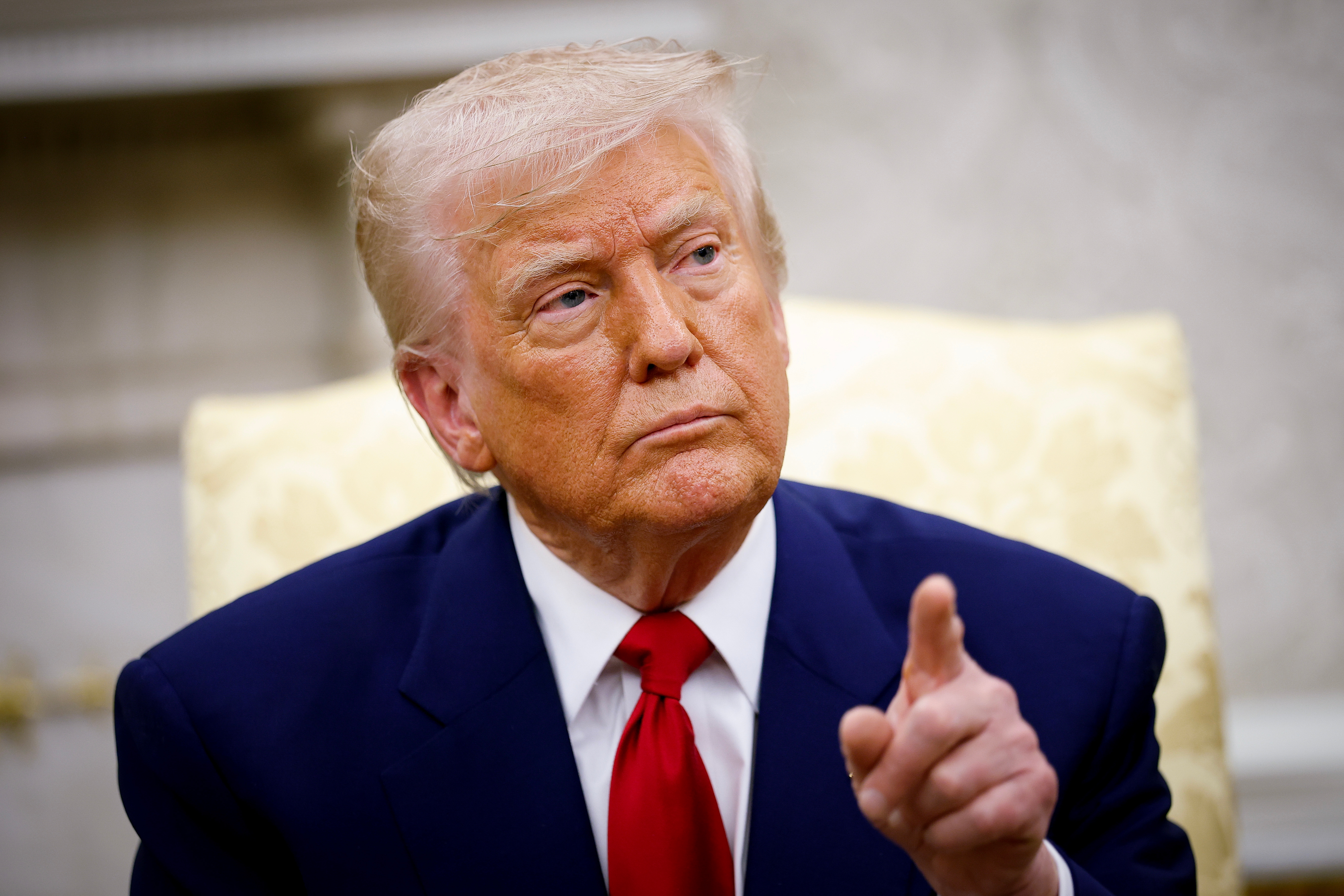 A person in a suit and tie is sitting, pointing forward with a serious expression