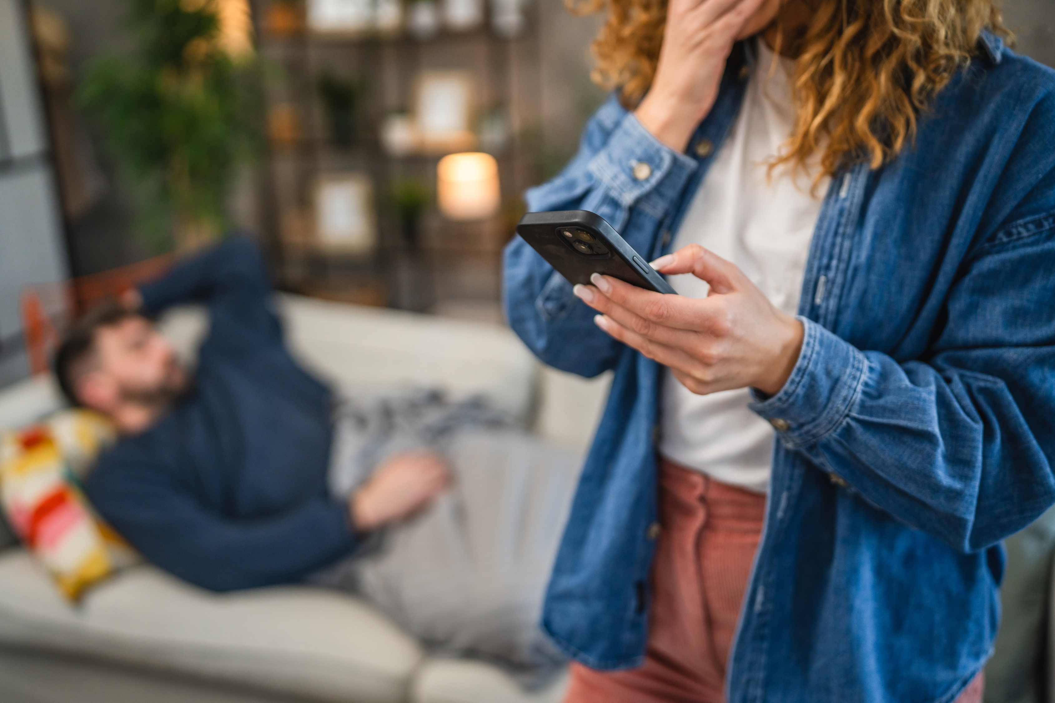 A person stands using a smartphone, looking serious, while another person relaxes on a couch in the background