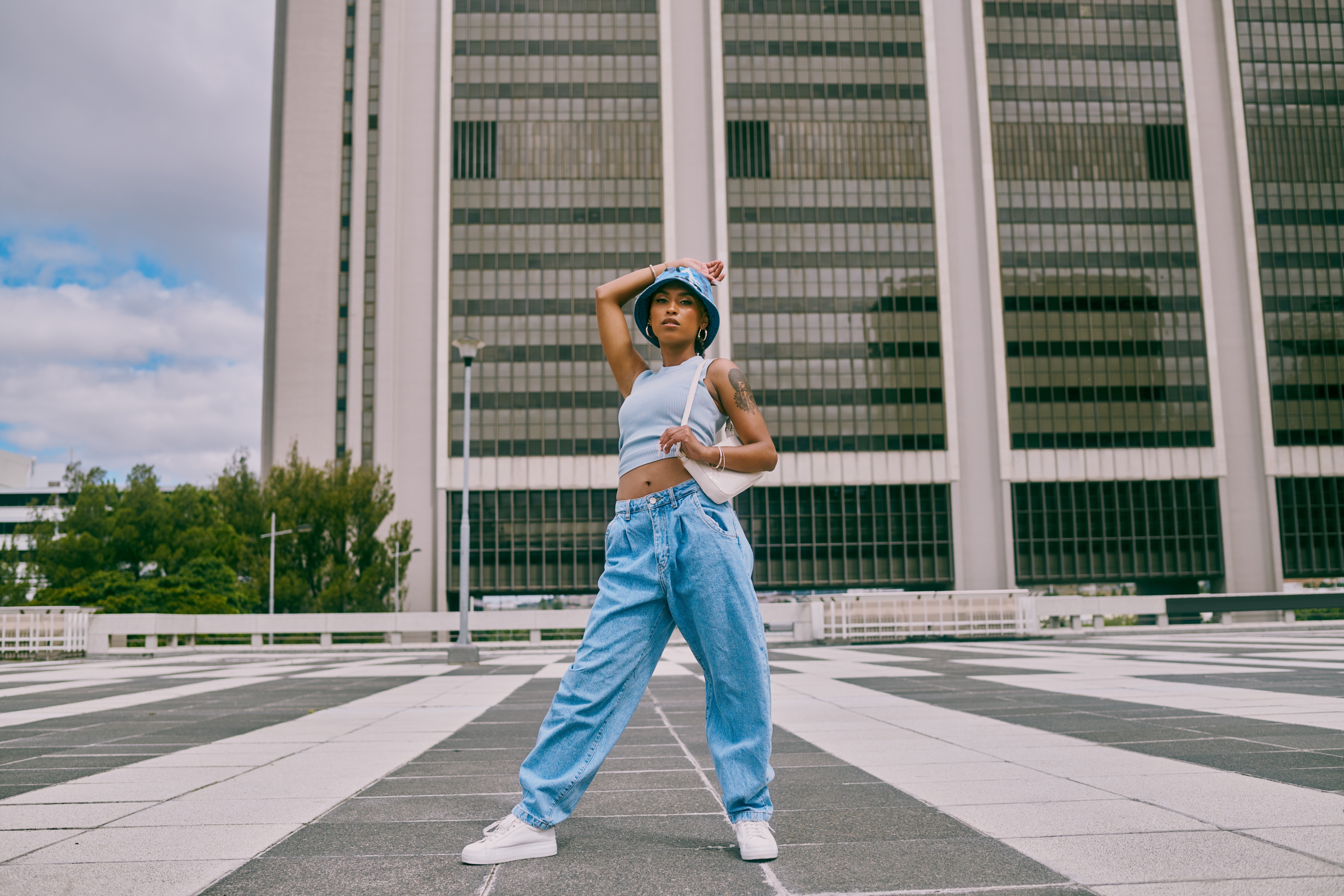 Person poses confidently outdoors wearing relaxed-fit jeans, a crop top, and a bucket hat, against a modern urban backdrop