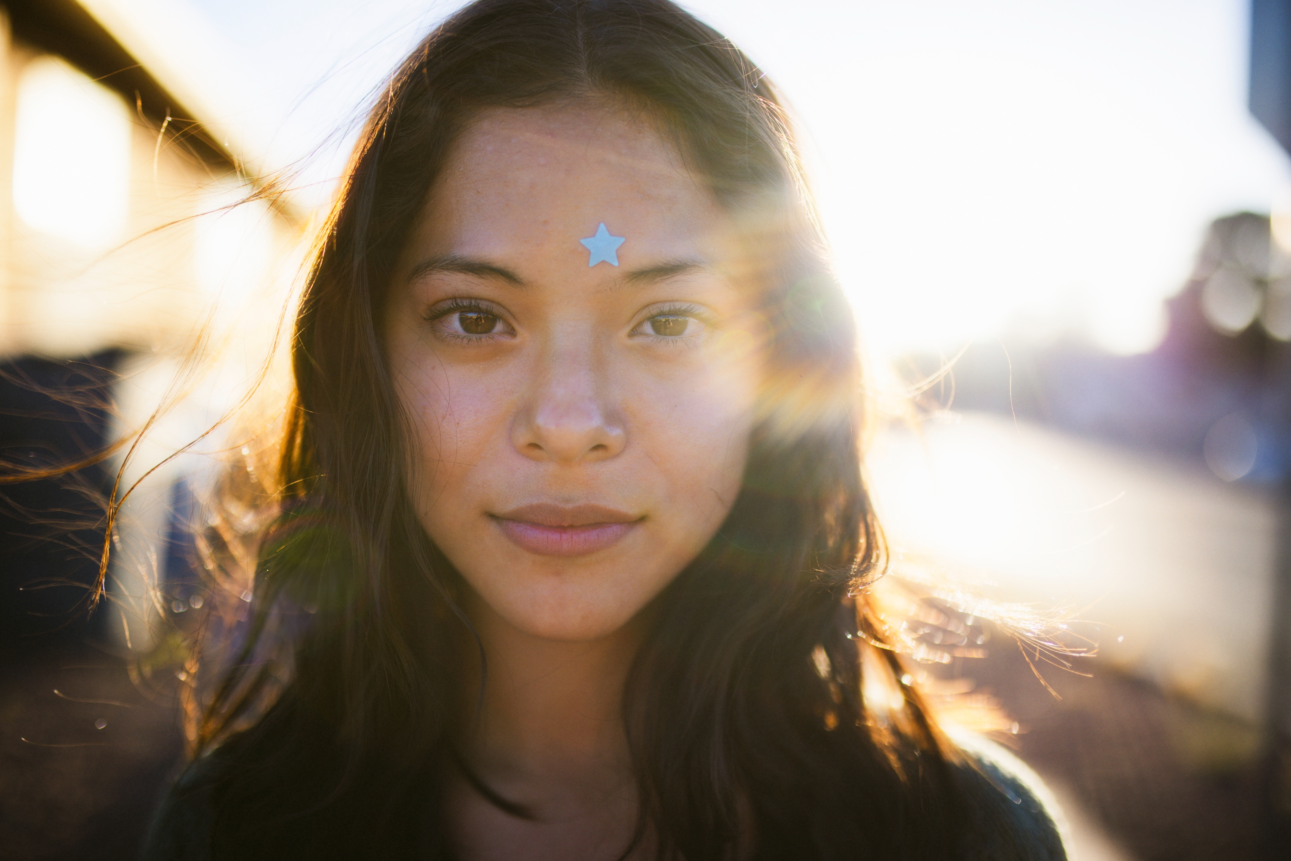 Person outdoors with sunlight behind, wearing a star sticker on forehead. Casual style, natural look