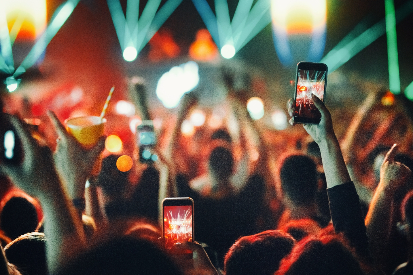 Audience at a lively concert, holding up phones to capture the performance on stage, with beams of light enhancing the vibrant atmosphere