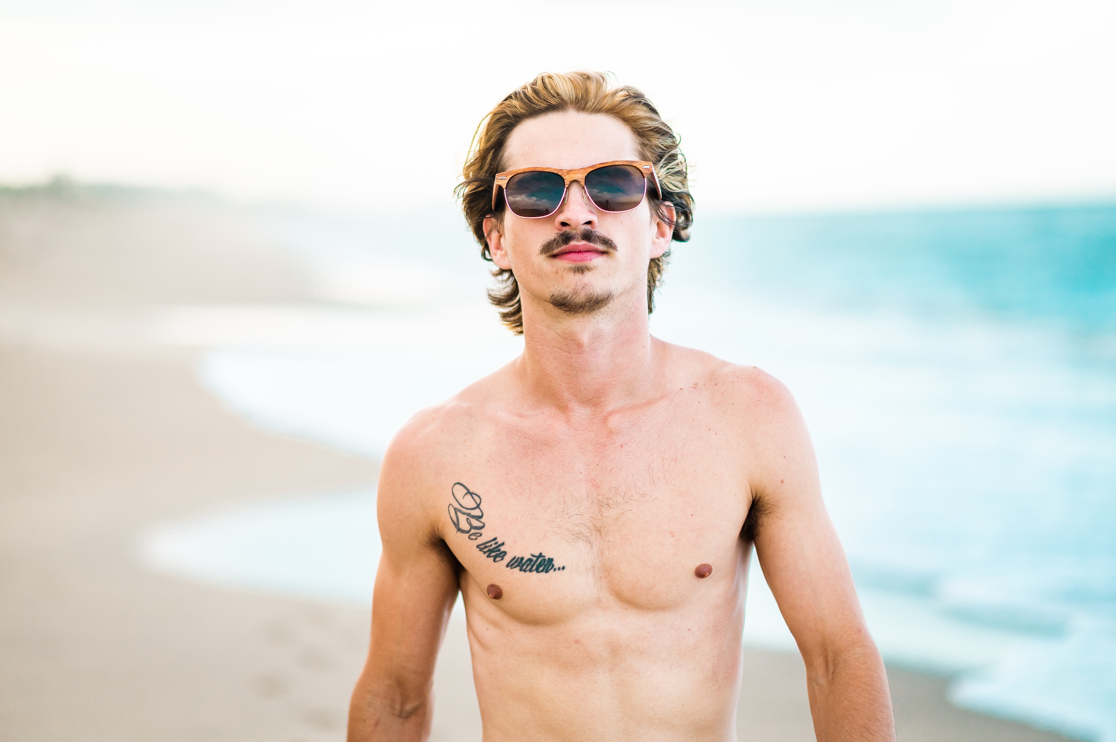 Man on the beach wearing sunglasses, with a tattoo on his chest, looking confidently at the camera. Waves are visible in the background